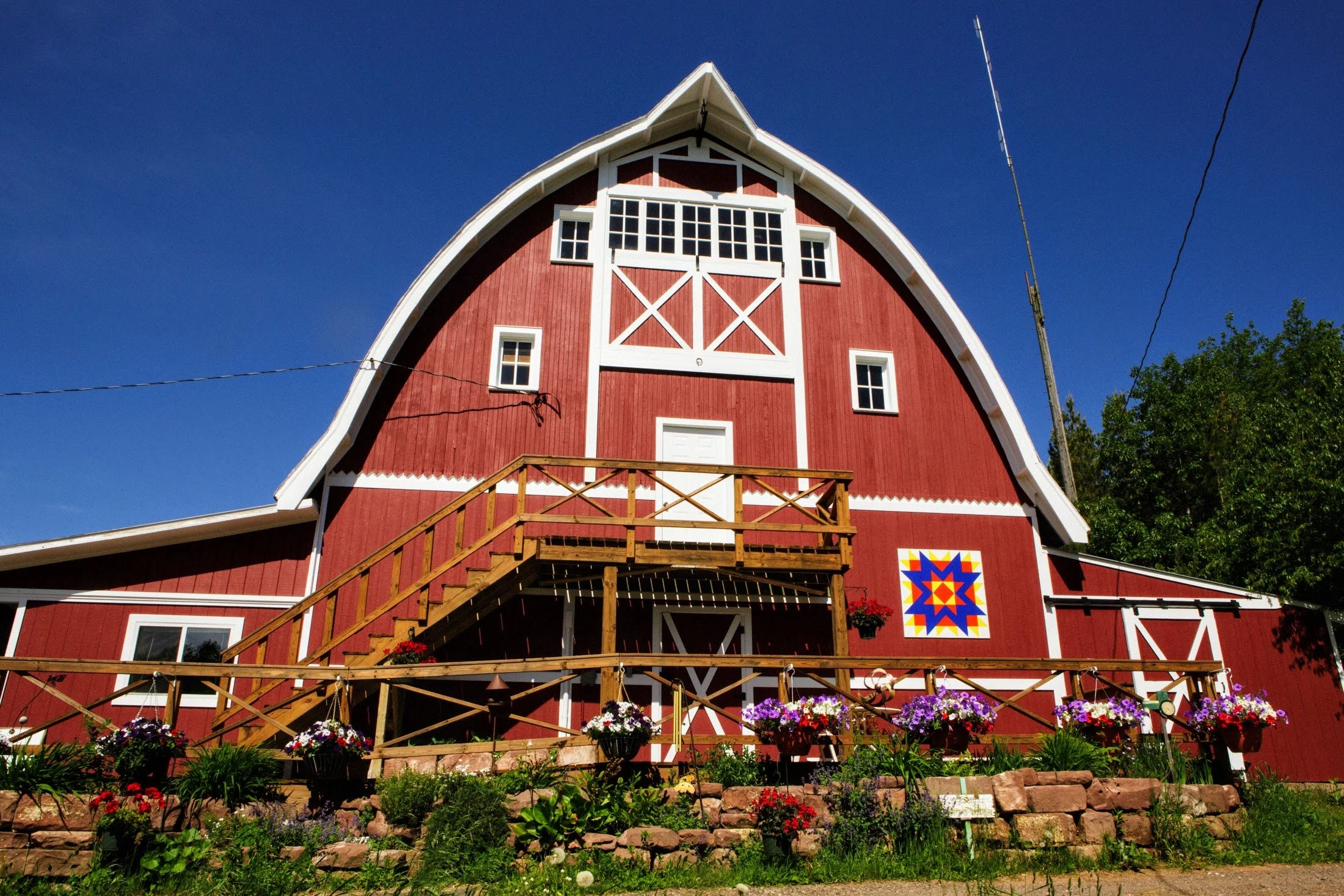 A large red barn with white trim, a wooden staircase leading to a door, several small windows, and a colorful quilt pattern on the side. There are flower boxes with blooming flowers and greenery in front of the barn, under a clear blue sky.