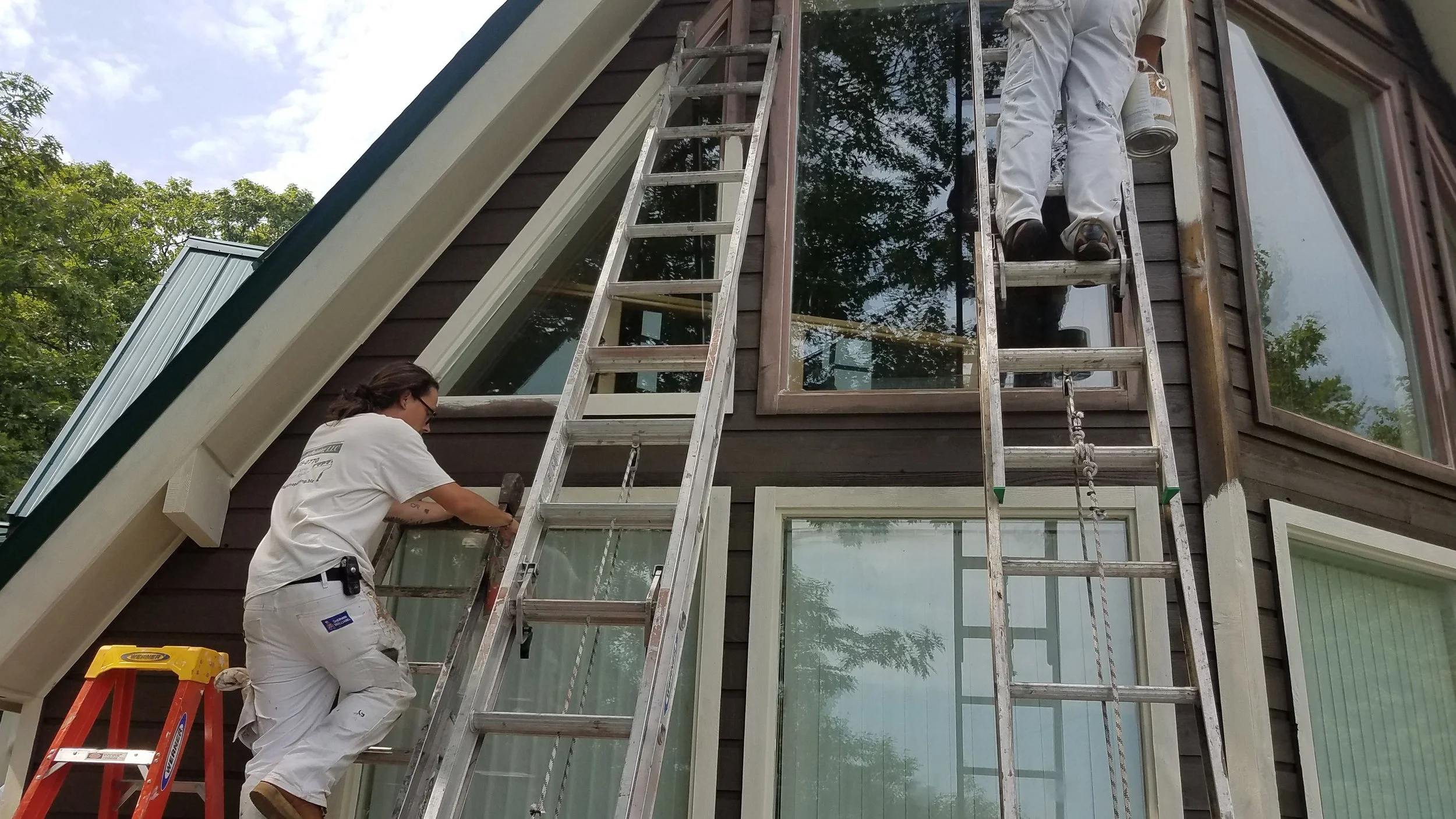 Two workers on laddersinstalling a large window in a two-story house with dark wooden siding and multiple windows, surrounded by green trees.