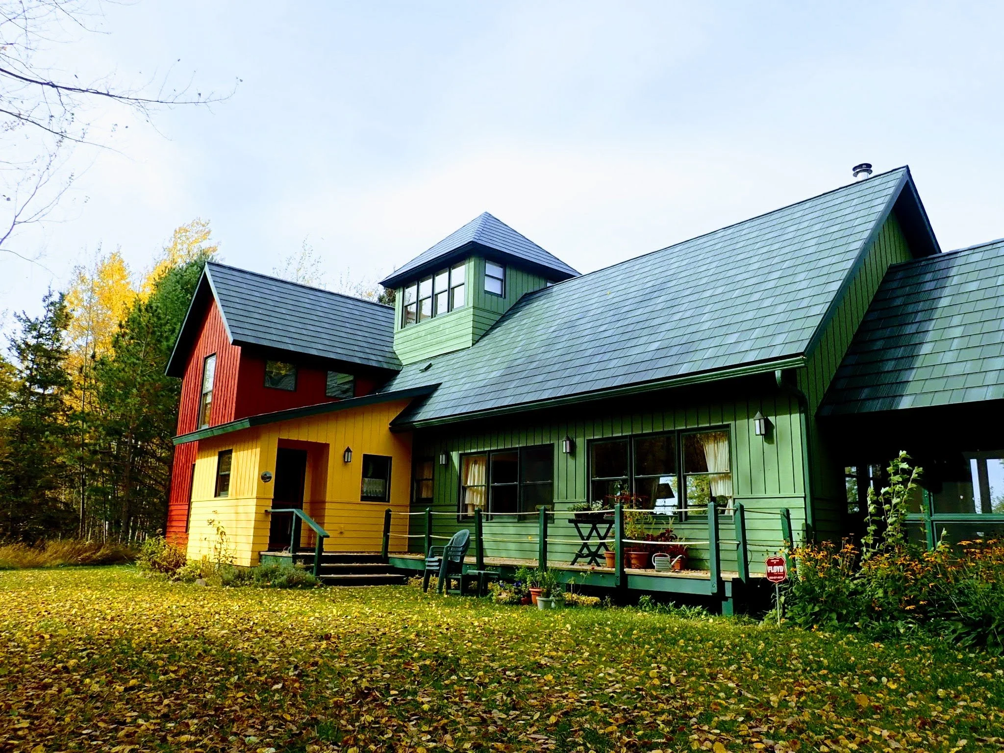 Colorful house with yellow, green, and red siding, multiple steep roofs, large windows, and a small porch in a yard with fallen leaves and surrounded by trees.