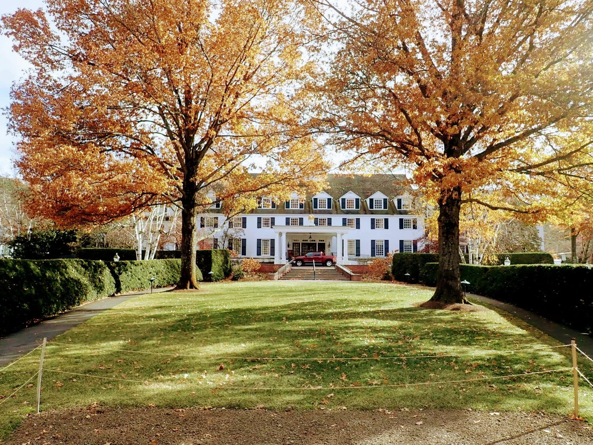A large white mansion with black shutters, surrounded by a manicured lawn and trees with orange and yellow fall foliage.