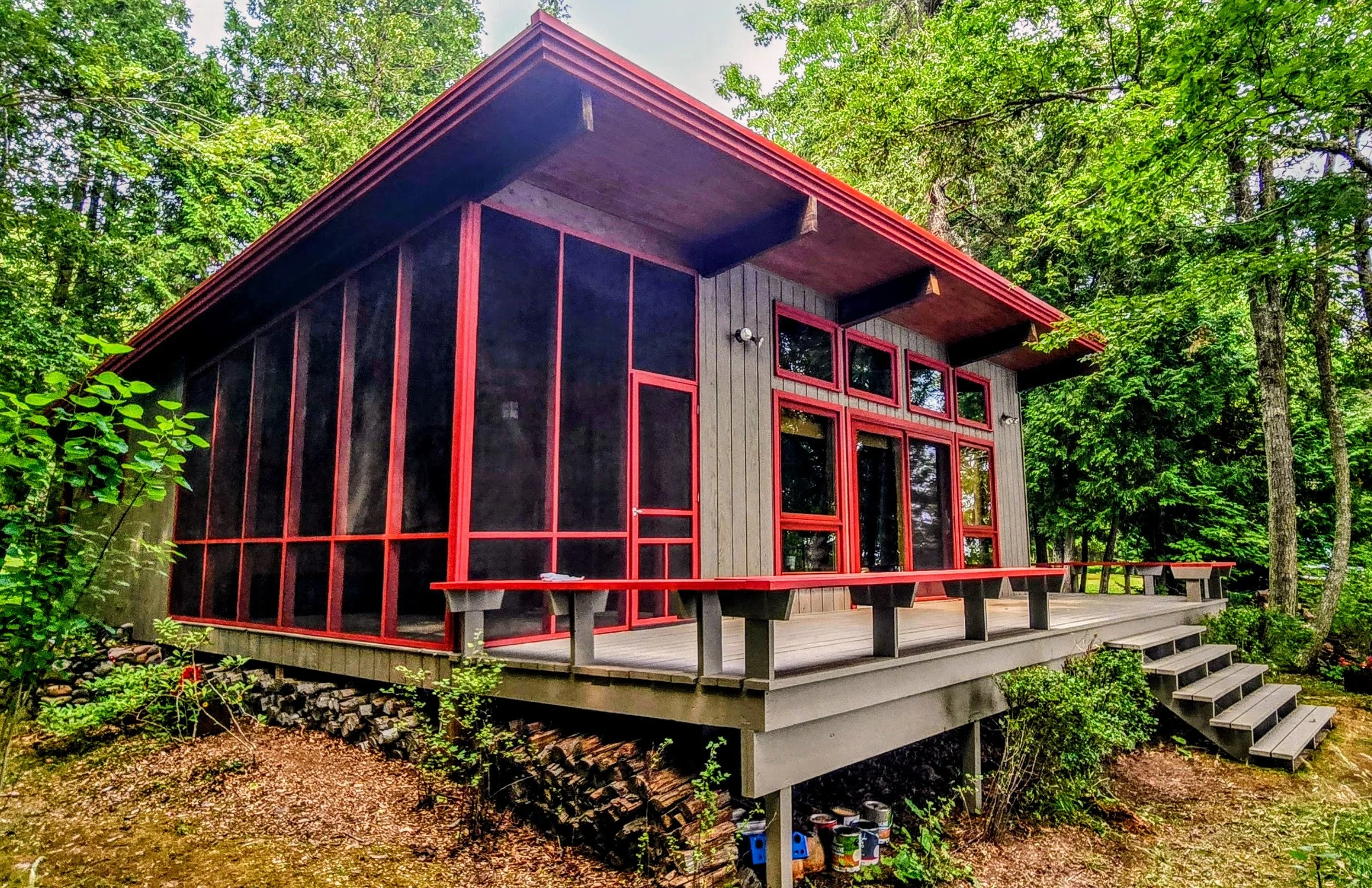 A modern wooden house with large red-framed windows and a screened-in porch, surrounded by trees and greenery.