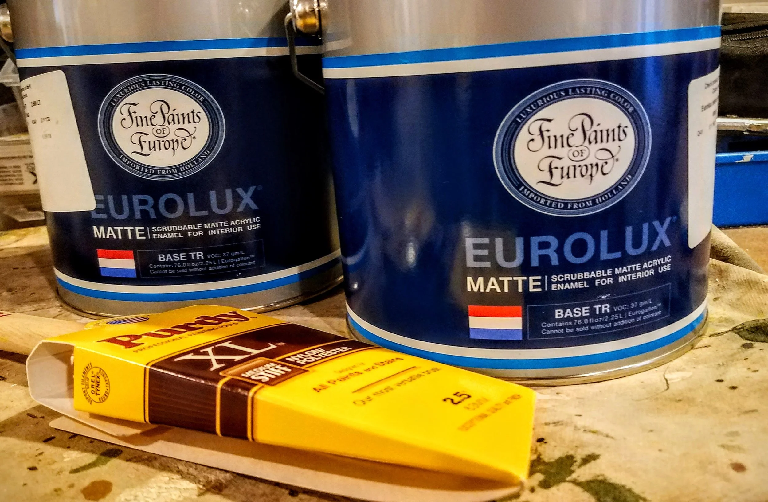 Two blue cans of EuroLUX matte interior enamel paint and a yellow tube of Golden Artist Colors paint on a wooden surface.