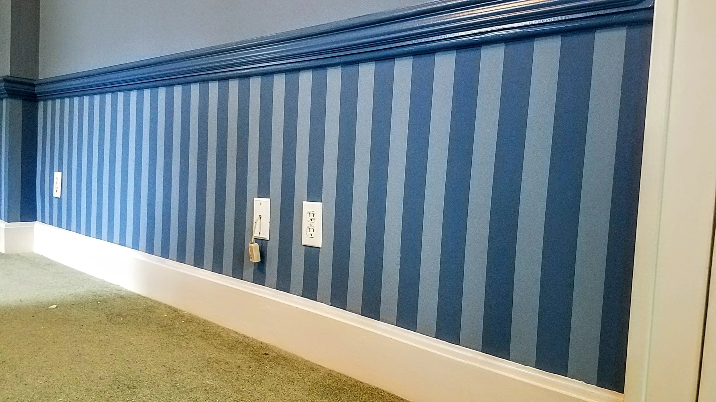 Blue striped wall with white electrical outlets and a telephone hanging from one outlet, beige carpet, and white baseboard.