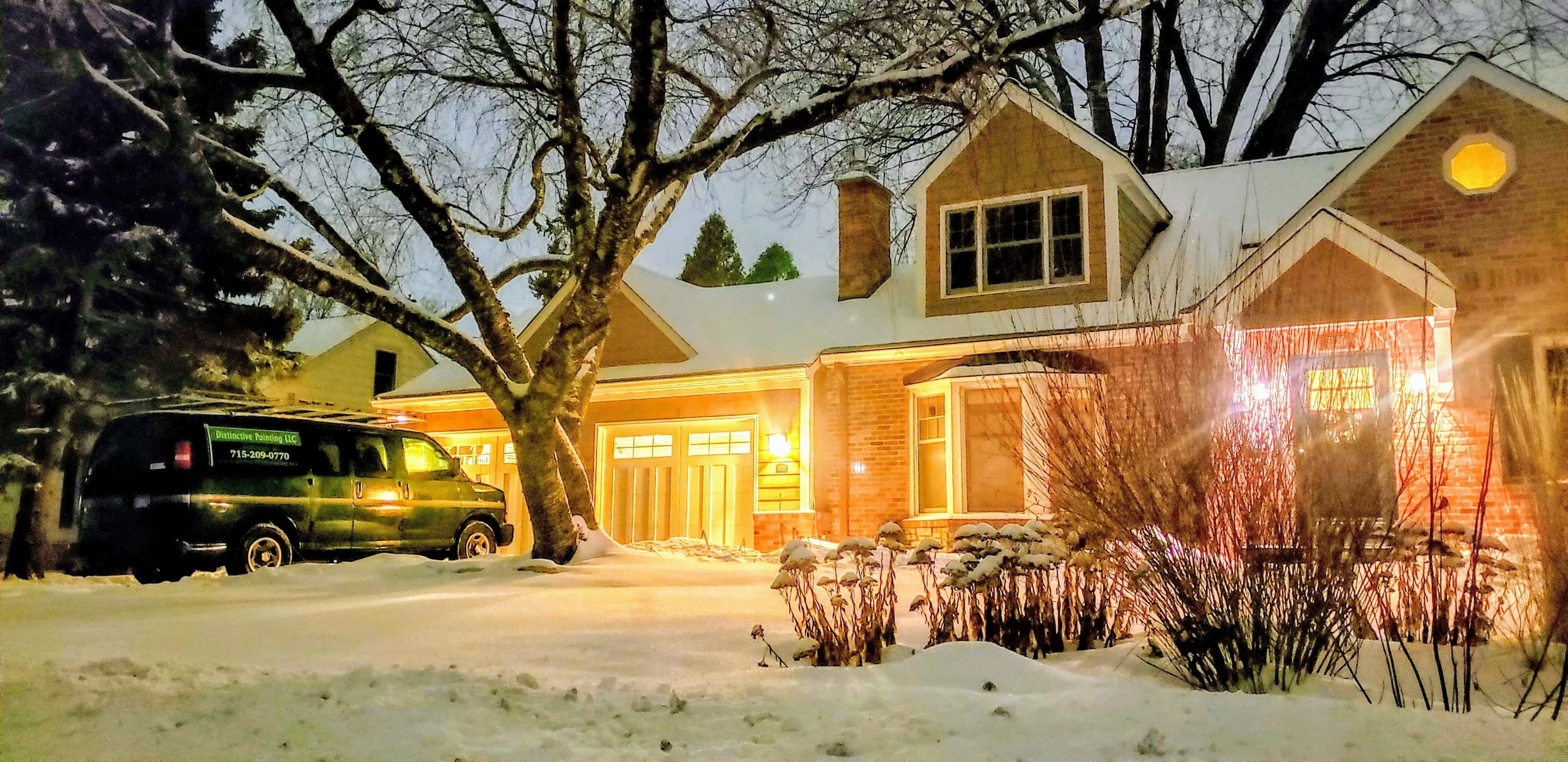 Snow-covered suburban house illuminated with warm lights at night, with a large tree in the front yard and a black van parked nearby.