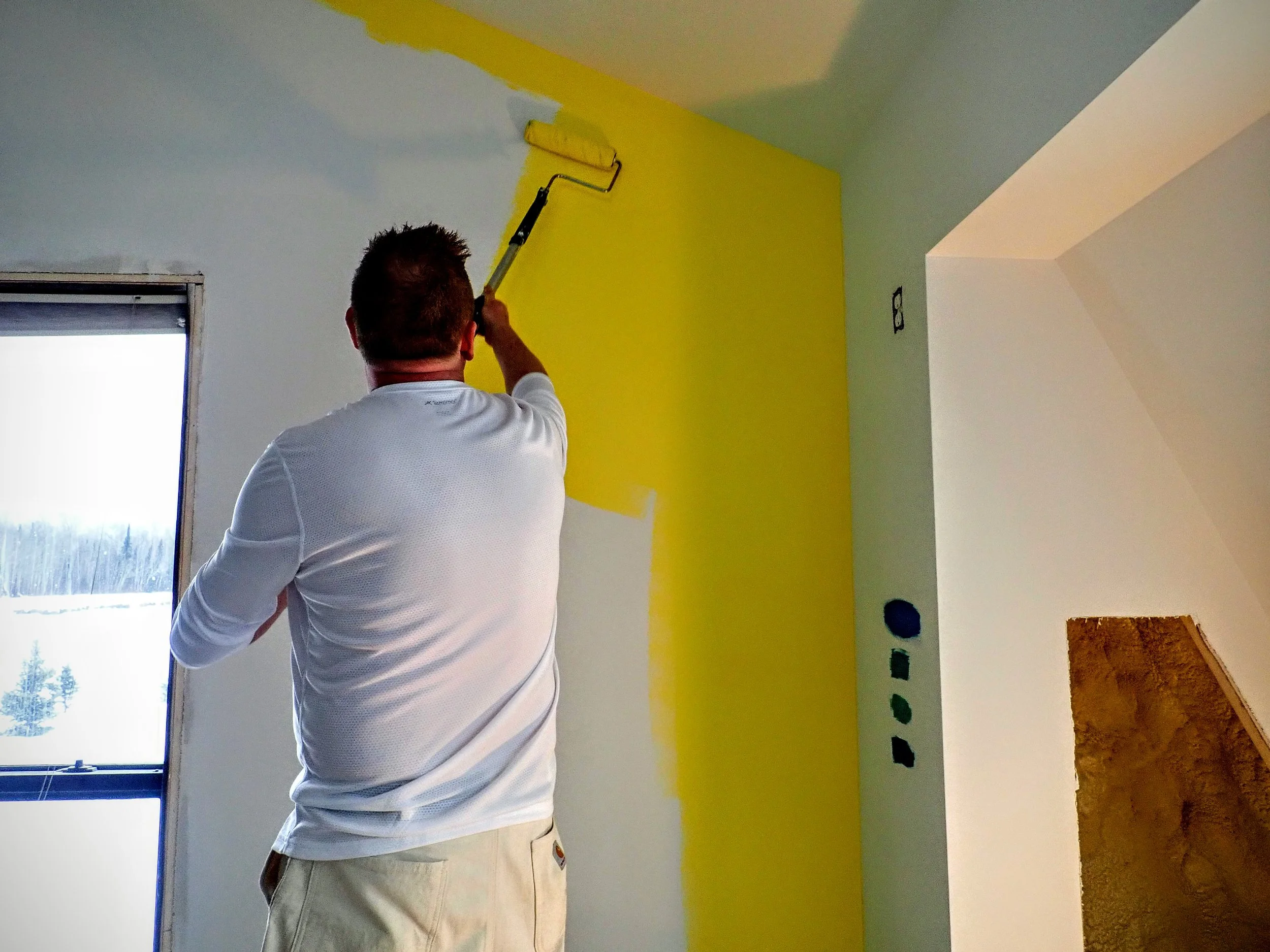 A person painting a wall yellow with a paint roller in a room with a large window.