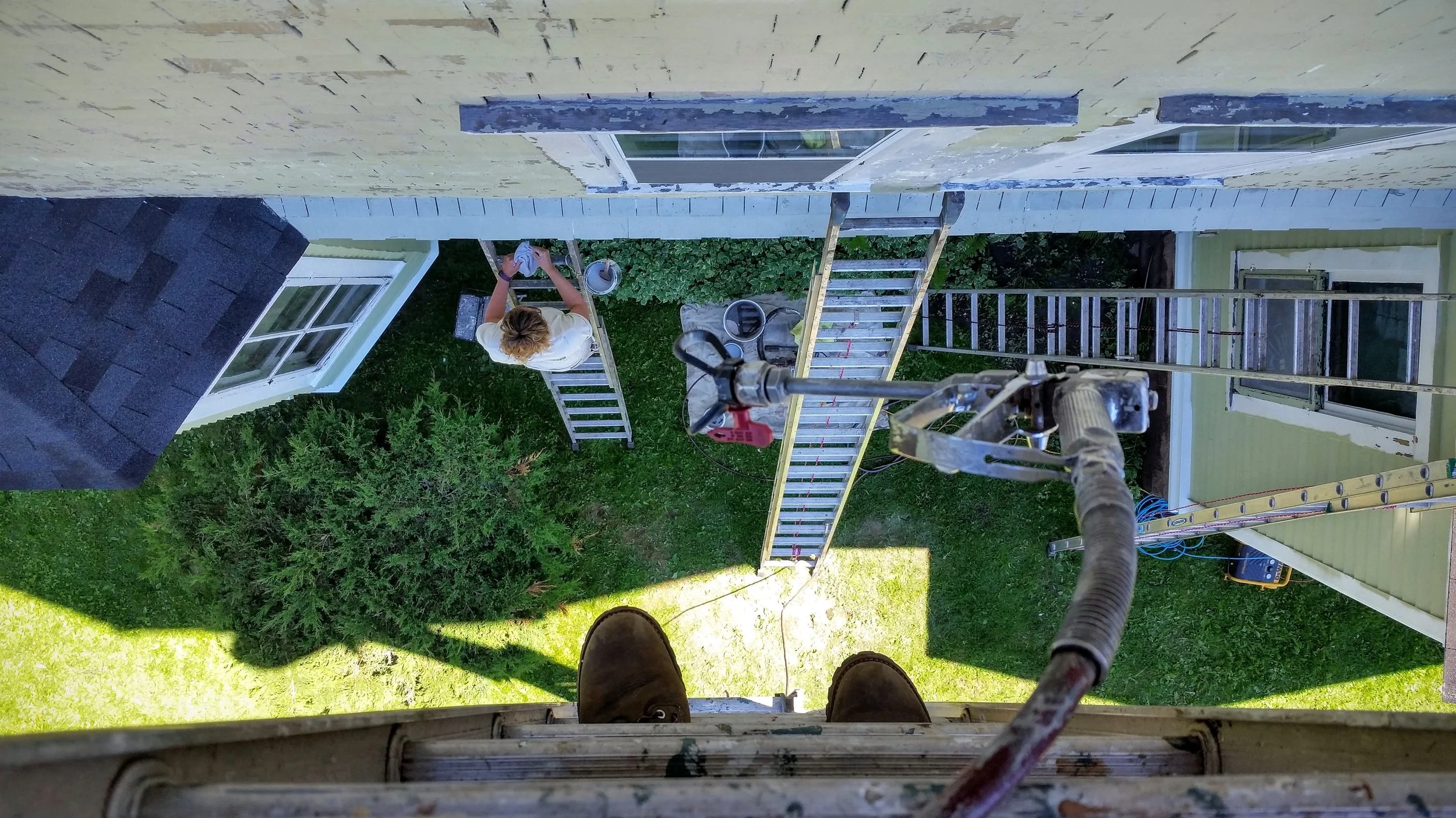 Person on a ladder cleaning the exterior of a house with scaffolding, shown from above, with gardening tools and a green yard below.