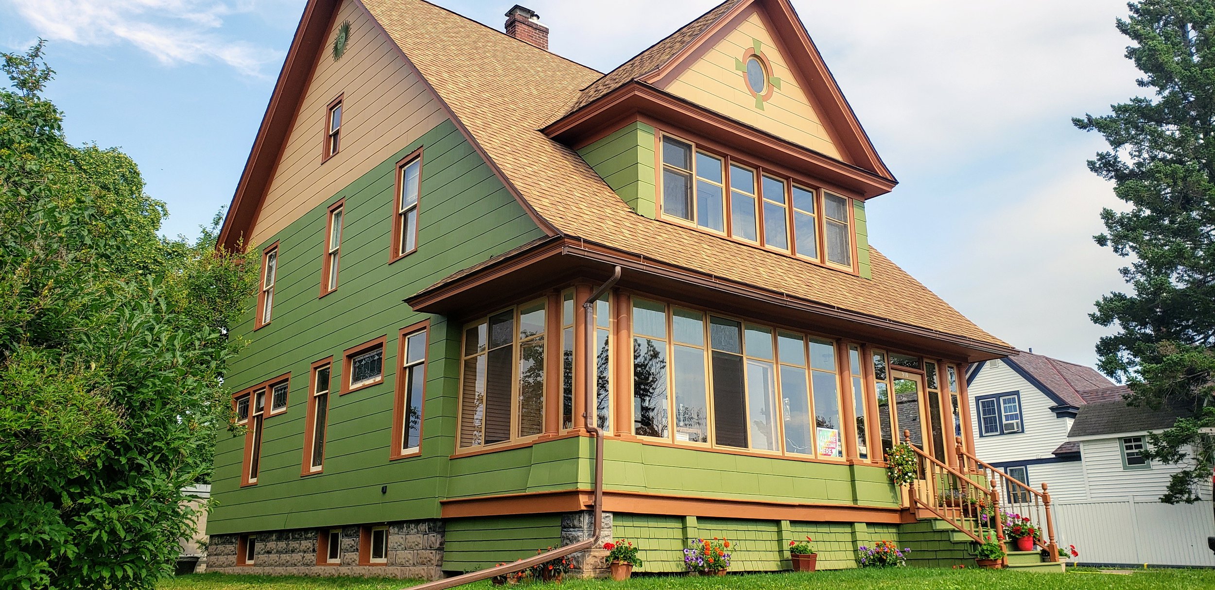 A colorful multi-story house with green and beige siding, large windows, and a front porch decorated with potted flowers. Surrounded by trees and a grassy yard.