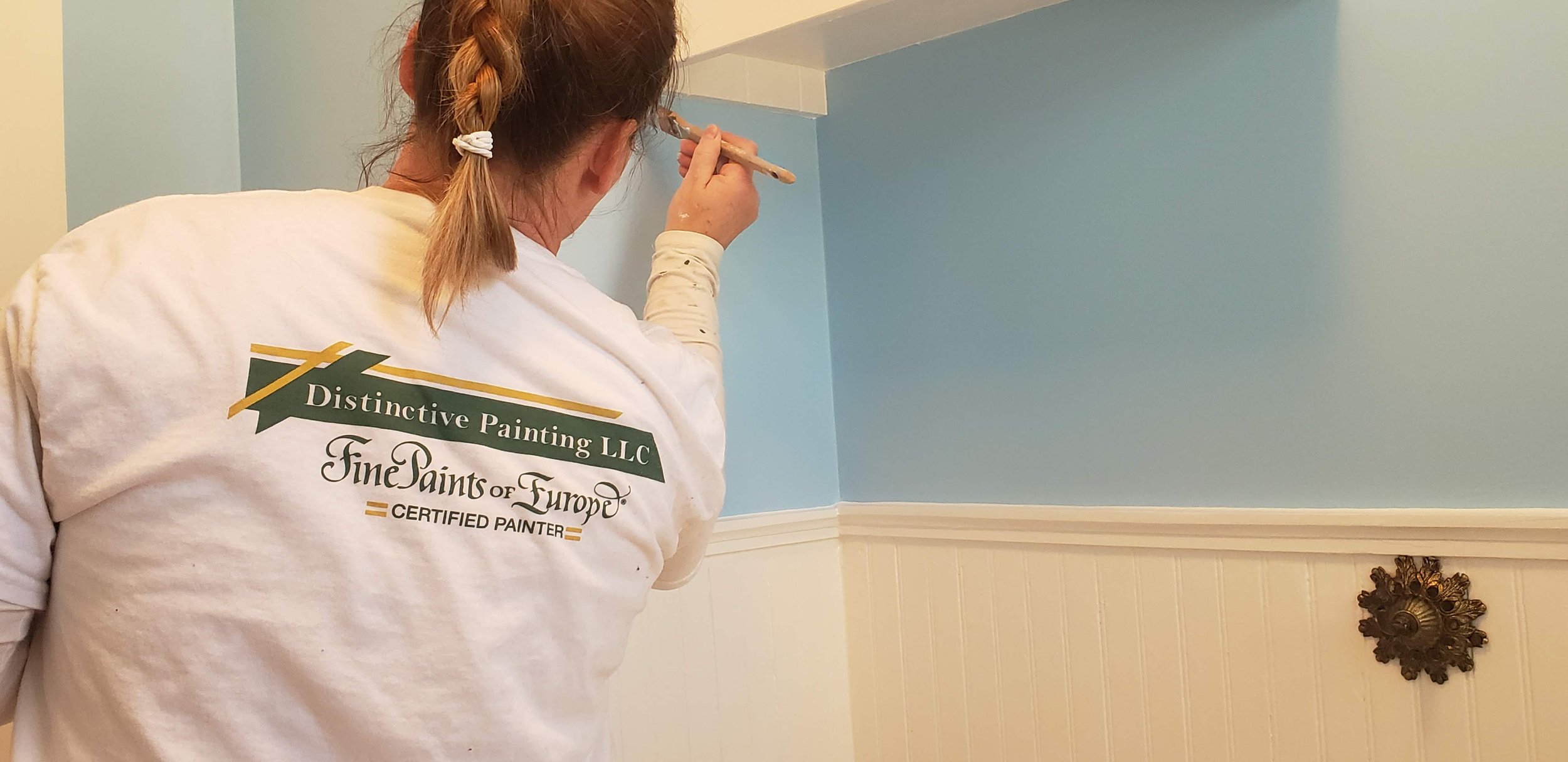 A woman painting a wall with a paintbrush, wearing a white shirt with the text 'Distinctive Painting LLC, FinePaints of Europe, Certified Painter' on the back, in a room with light blue upper walls and white lower paneling.