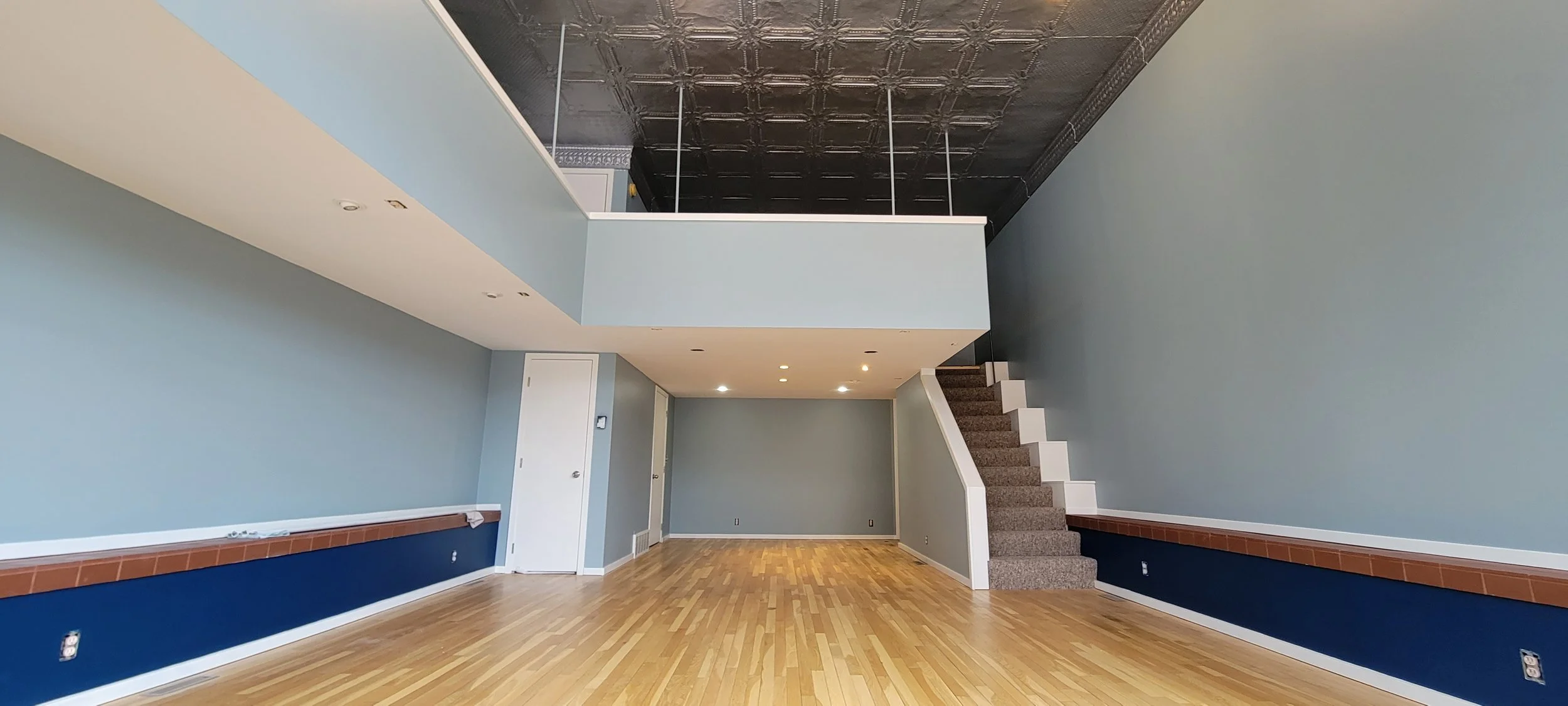 Empty room with hardwood floors, light blue walls, a stairway with brown carpet, and a second-floor balcony with decorative tin ceiling under construction.