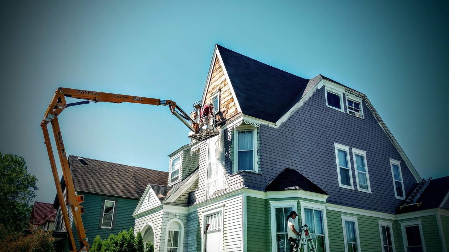 Workers on a lift working on the exterior of a multi-story house, which has different colored siding and a steeply pitched roof, during daytime with a clear sky.