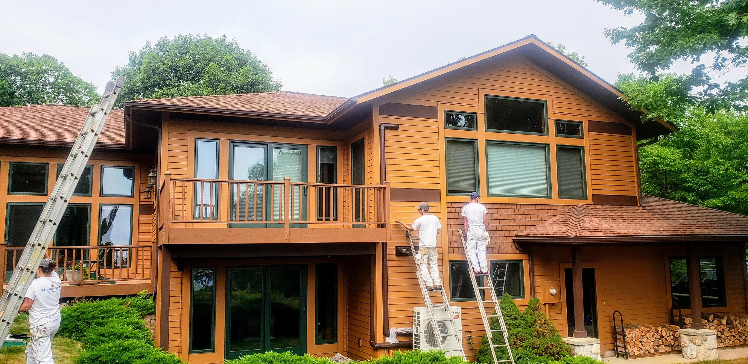 Workers painting the exterior of a large wooden house with multiple windows and a balcony, surrounded by trees and greenery.