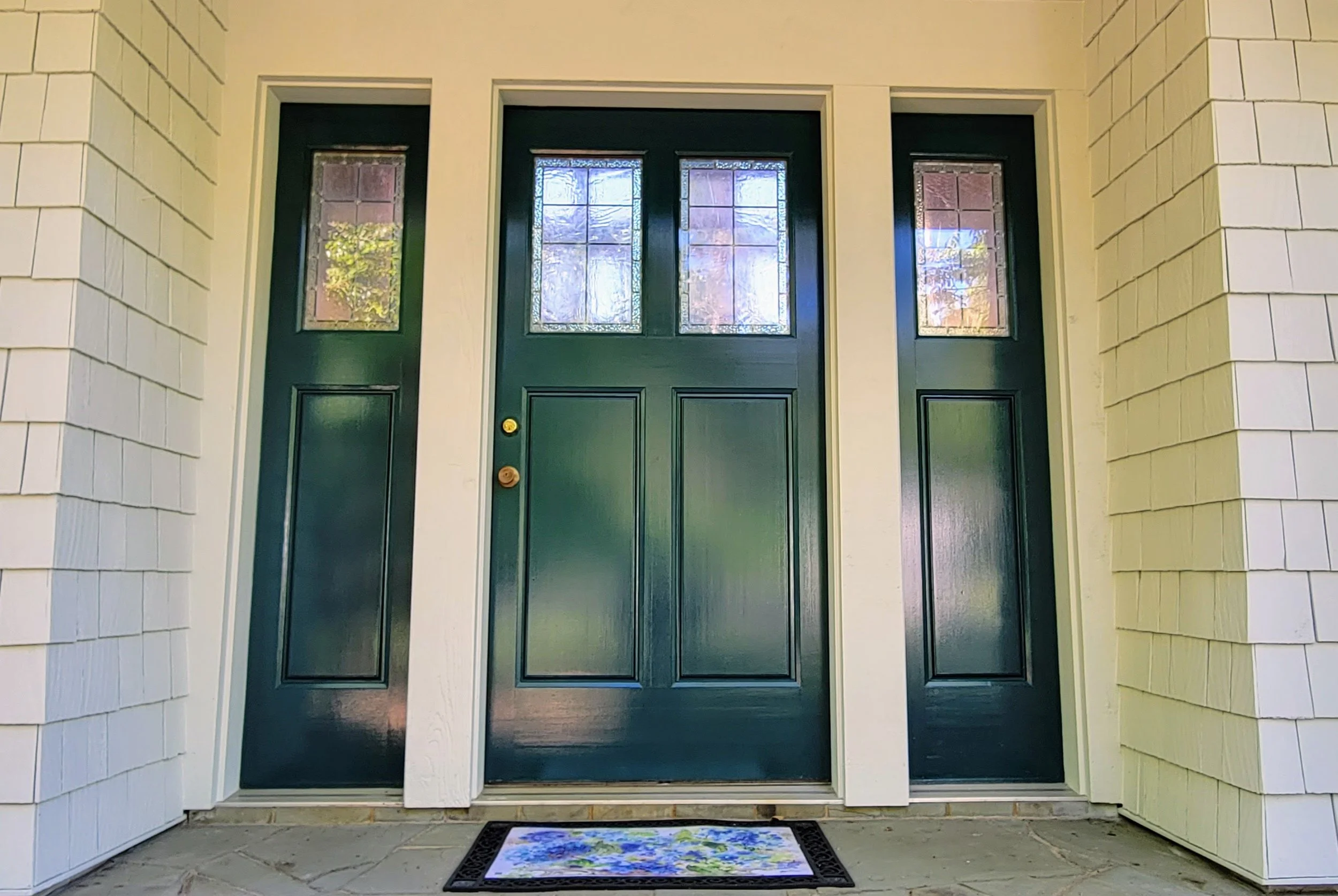 Front door with two smaller side doors, painted dark green, with windows at the top, beige house siding, and a floral welcome mat on the porch.