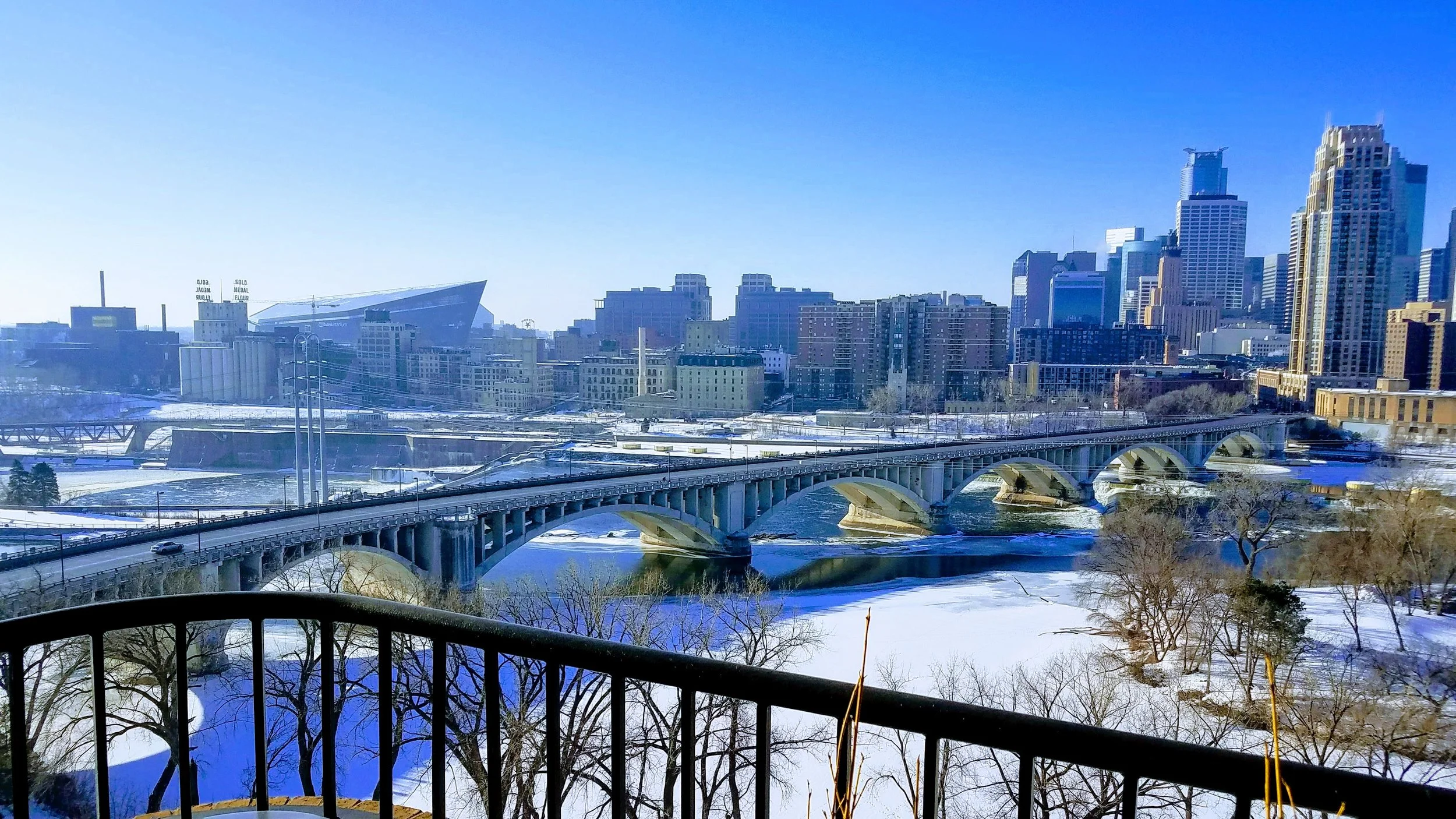 City skyline with tall buildings and a bridge over a frozen river, snow-covered trees and pathways in the foreground on a sunny winter day.