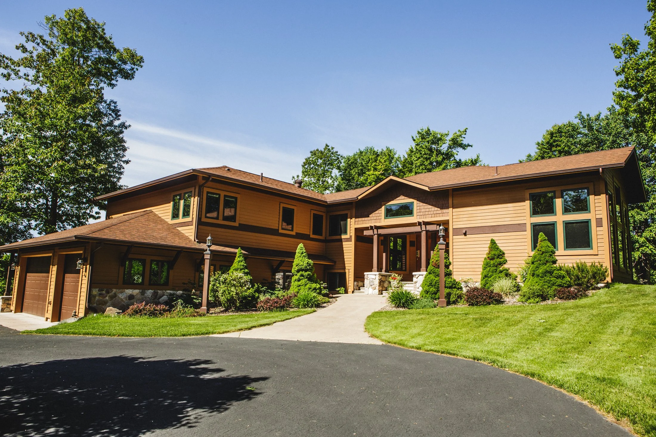 A large, modern house with multiple sections, brown exterior, surrounded by green trees and a well-maintained lawn, with a paved driveway leading to the garage.