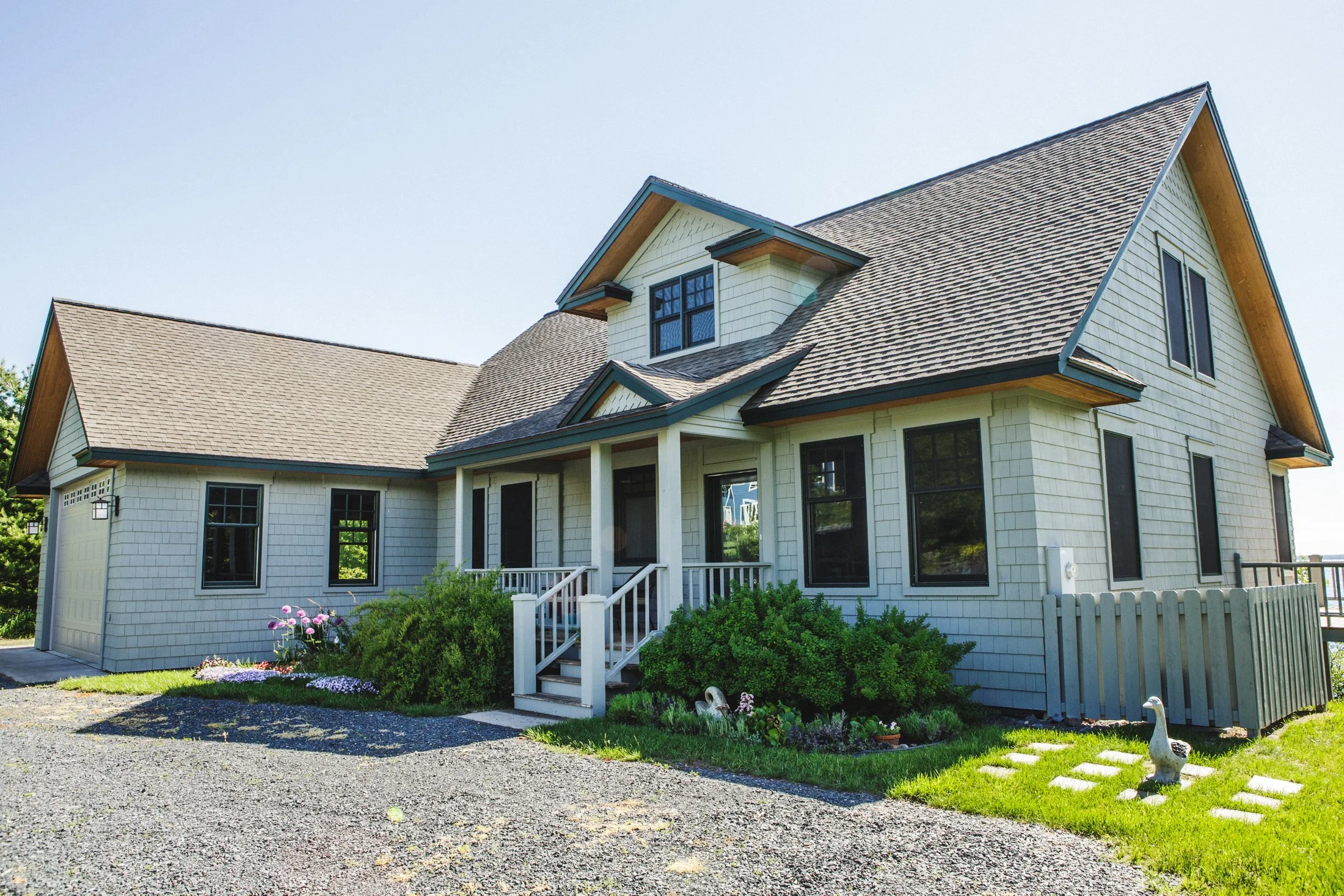A modern two-story house with gray shingles, black window frames, green bushes, and a small front porch with stairs.