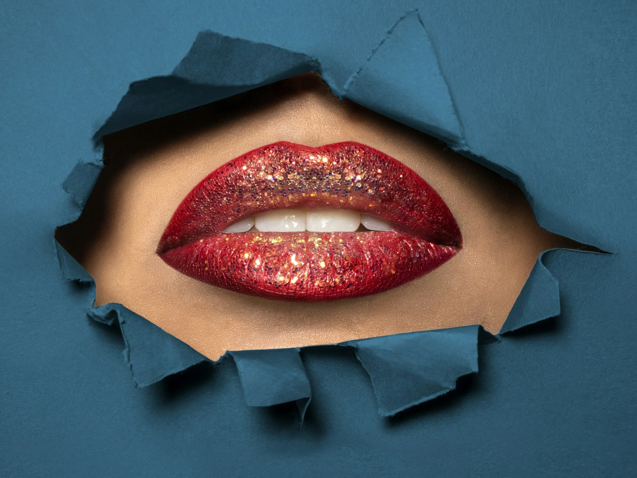 Close-up of lips with red glitter lipstick peeking through a torn piece of blue paper.