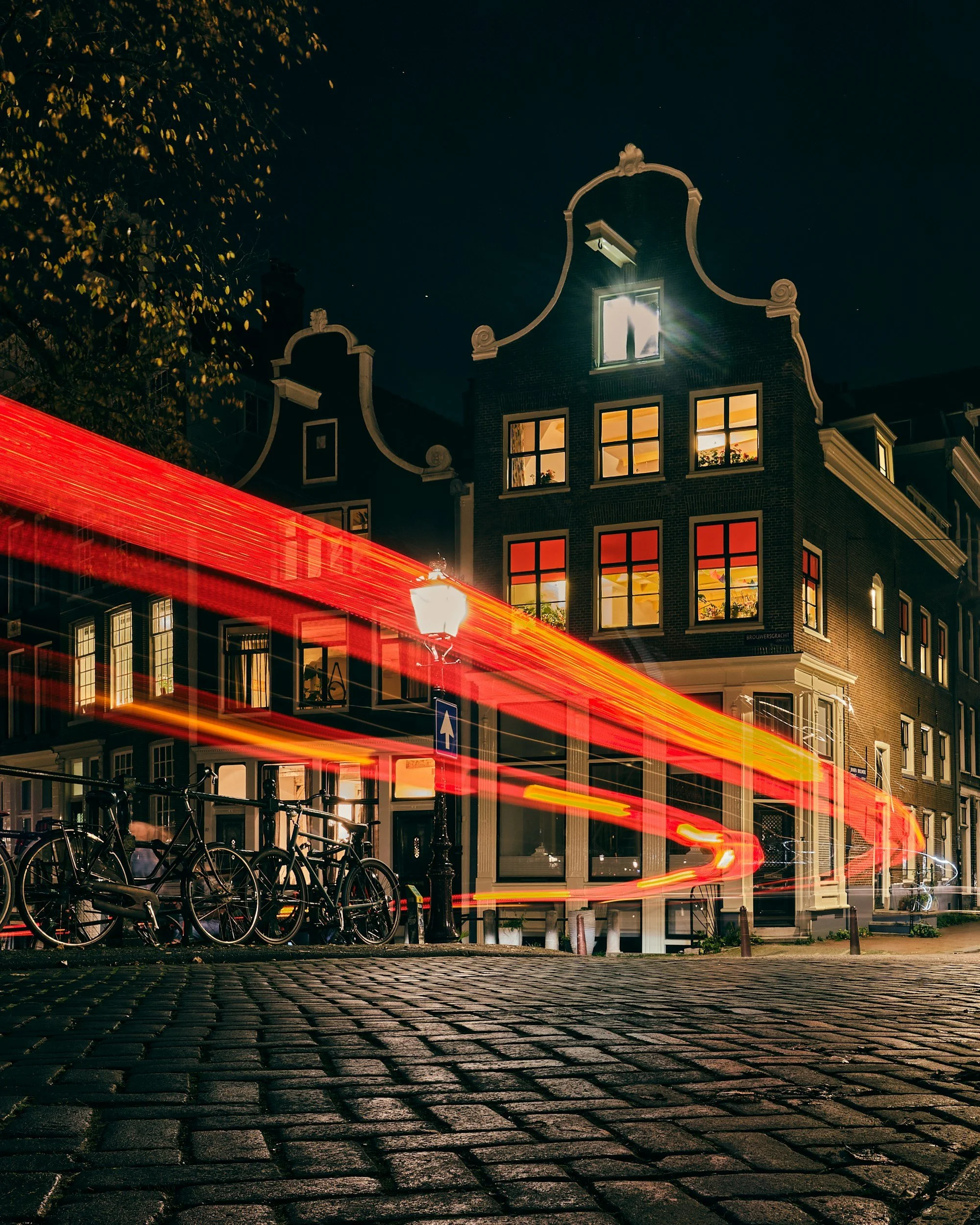 Nighttime scene in a city with historic buildings and bicycles parked along a cobblestone street. Light trails from passing vehicles streak across the image, and warm interior lighting is visible through the windows of the buildings.