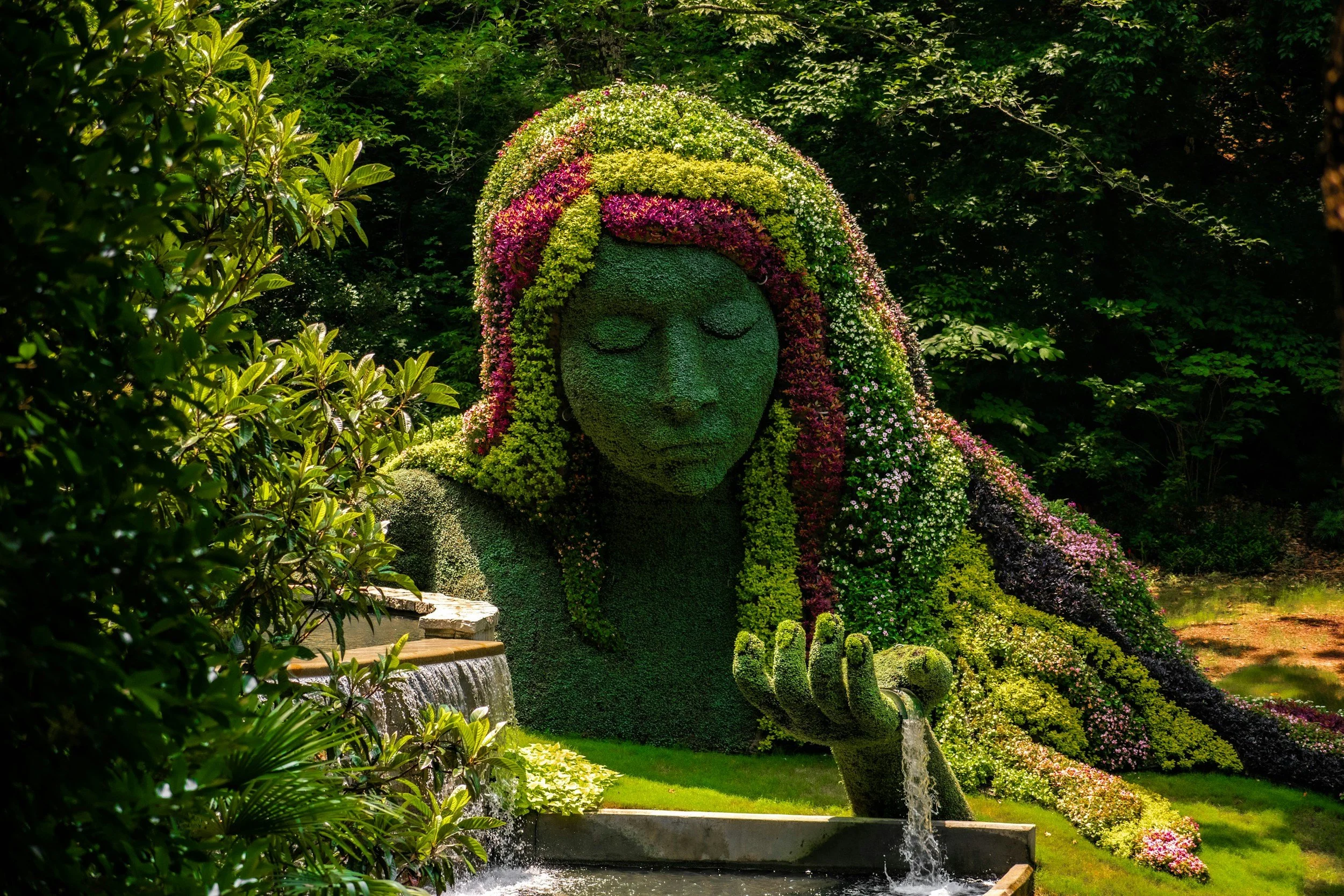 A large floral sculpture of a woman's face and hand, made from various colorful flowers and greenery, in a garden setting with trees and water features.