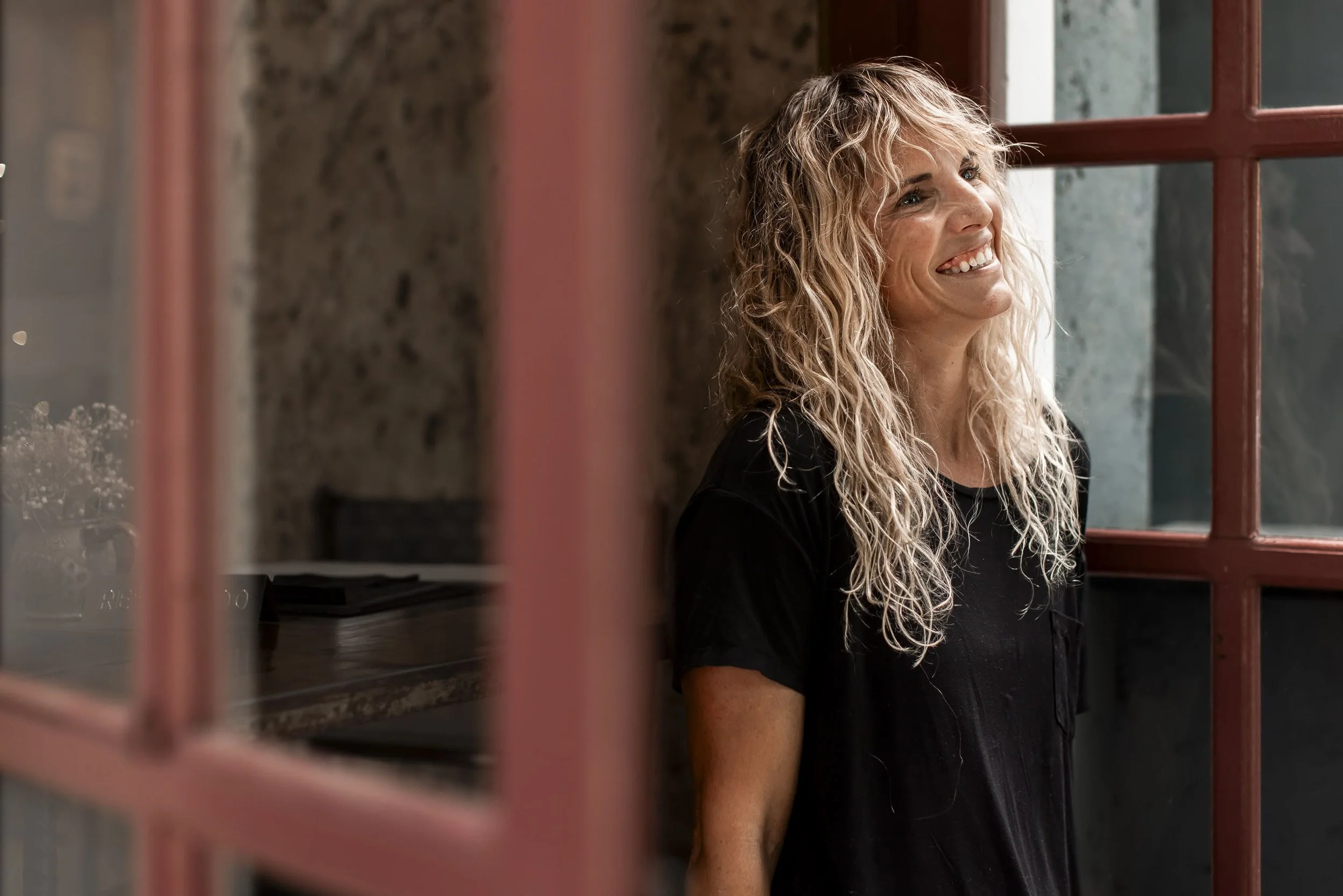 A woman with curly blonde hair smiling and looking out of a window with red framing.