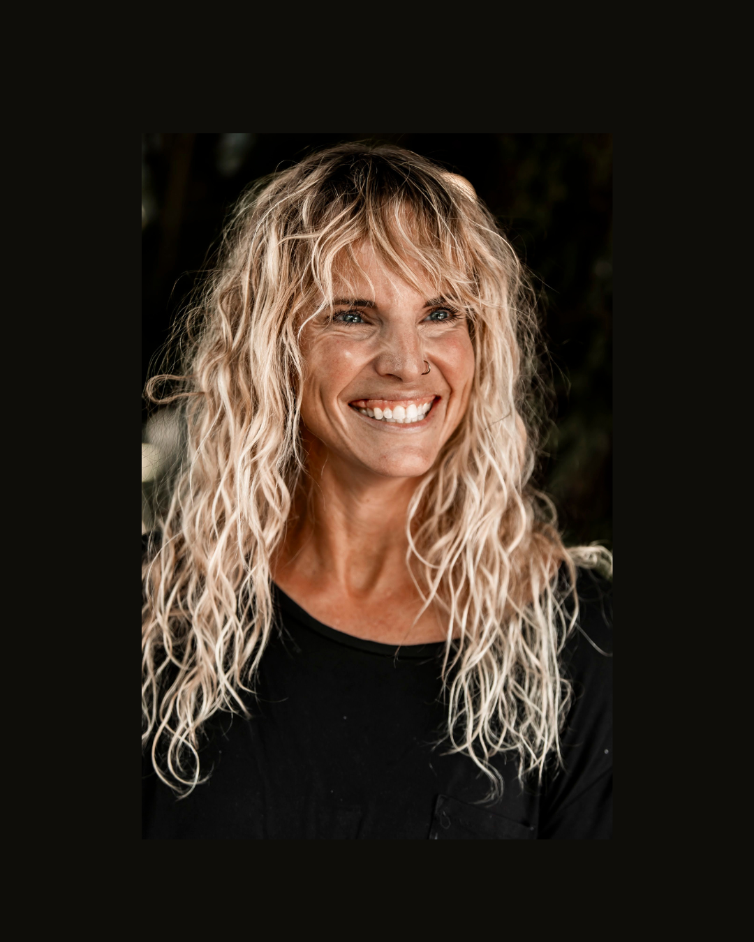 A smiling woman with long, curly blonde hair and a nose ring, wearing a black shirt, outdoors with a blurred natural background.