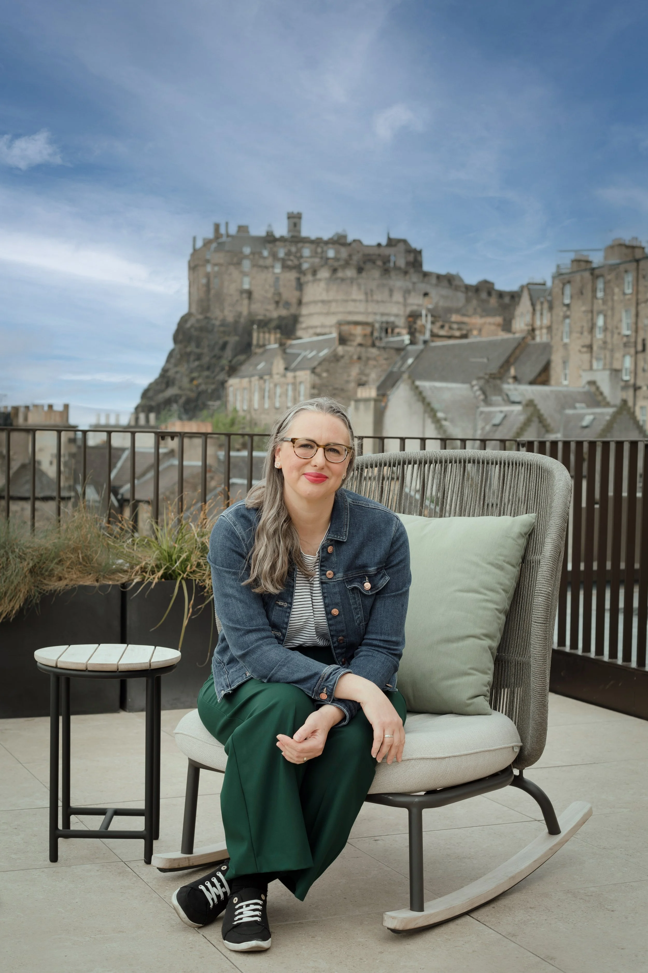 A woman with glasses and long brown hair sitting on a cushioned outdoor chair on a rooftop terrace with Edinburgh castle on a hill in the background.