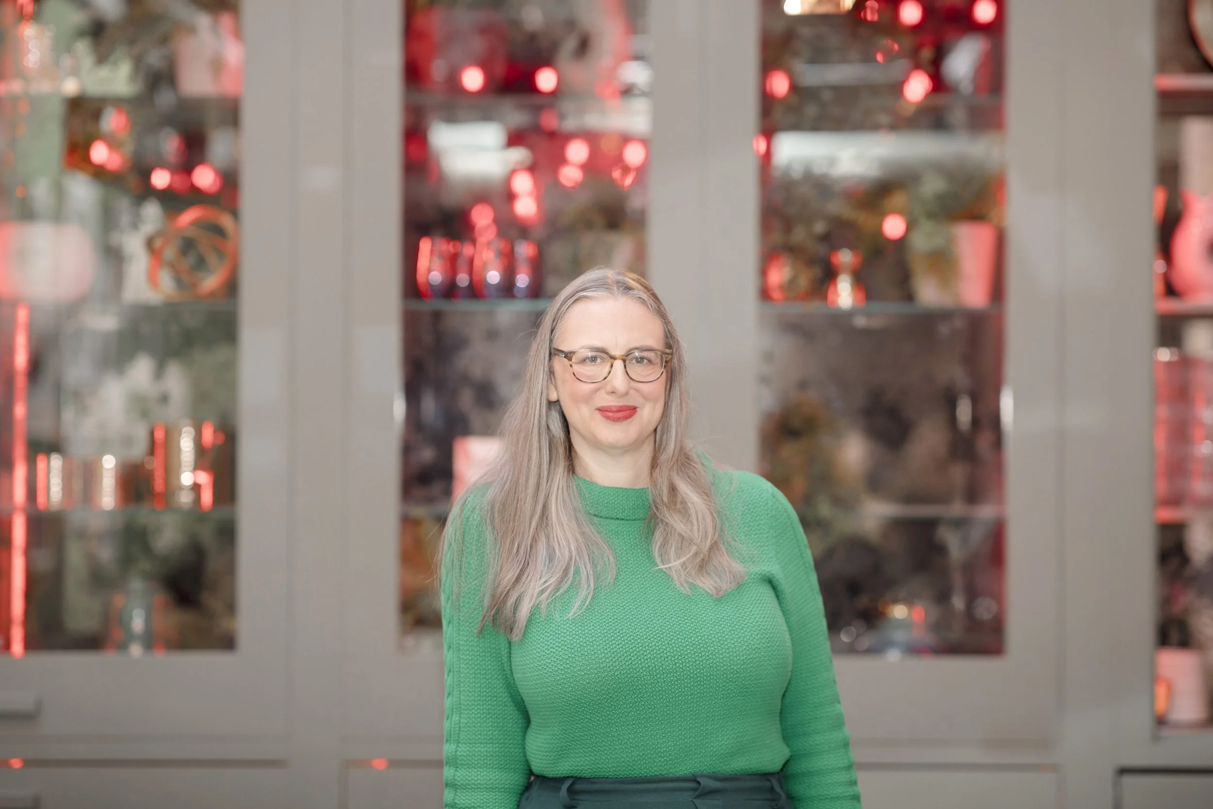 A woman with long brown hair and glasses standing in front of glass cabinets filled with decorative items, wearing a green sweater.