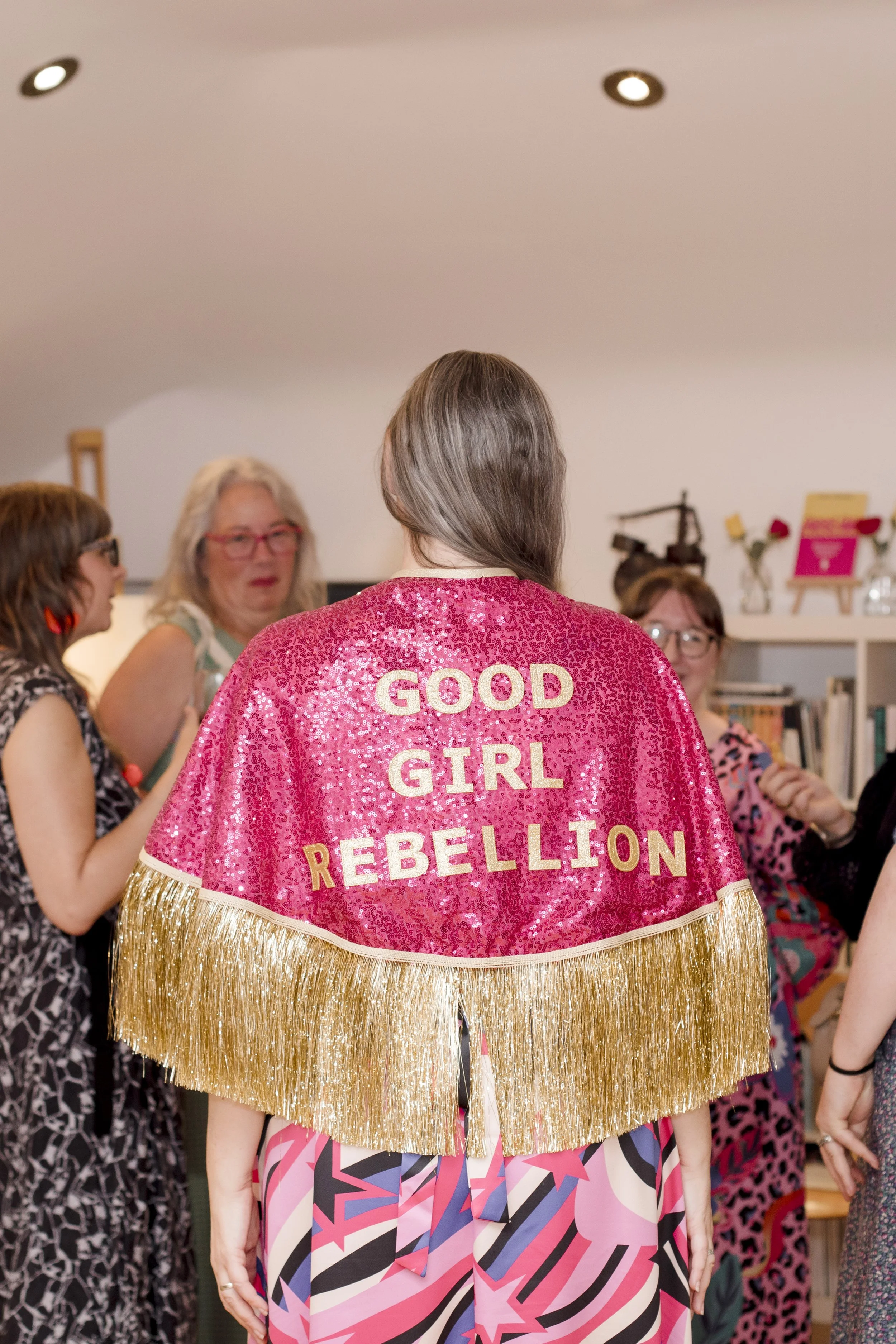 Photo of the back of a woman wearing a pink sequin cape with good girl rebellion on in gold lettering