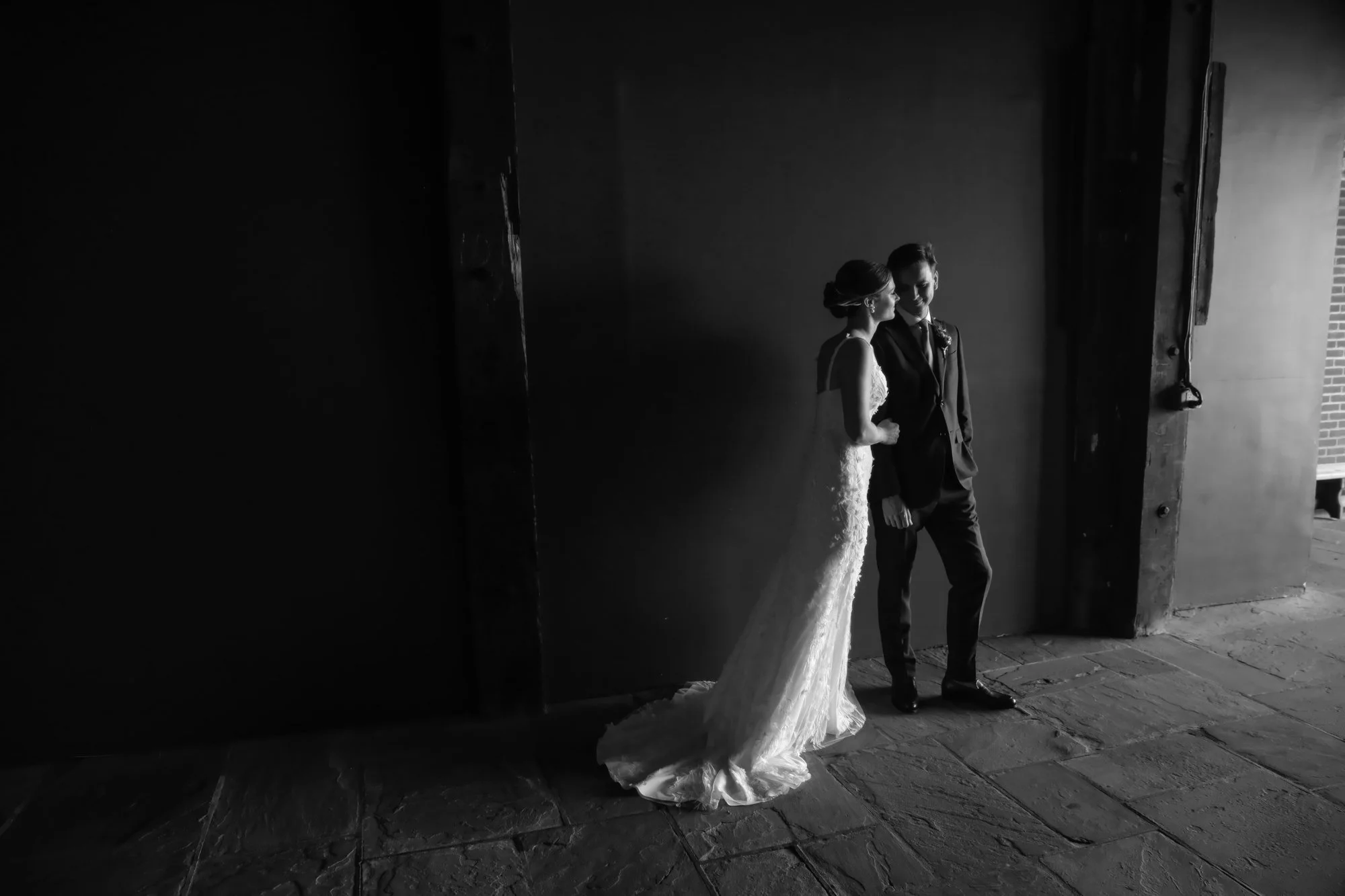 Black and white photo of a bride and groom standing close together in an industrial-looking area, with the bride in a wedding gown and the groom in a suit, looking at each other.