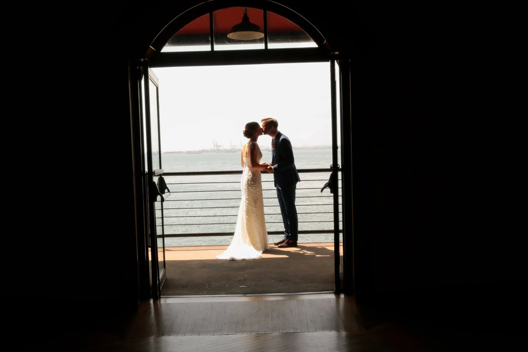 A bride and groom share a kiss while holding hands on a balcony overlooking the water, seen through an arched doorway with open doors, during sunset.