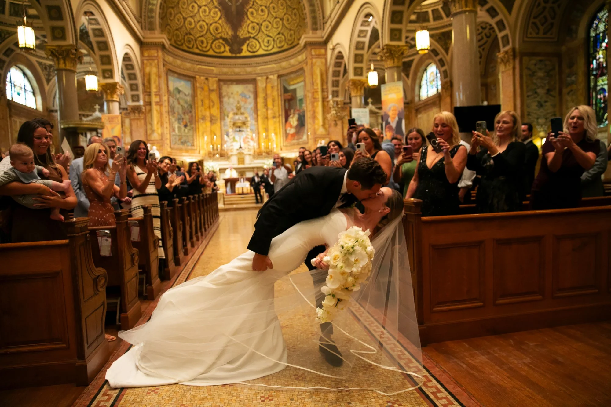 A bride and groom sharing a kiss in a church during their wedding ceremony, with guests watching and taking photos, in a grand decorated church interior.