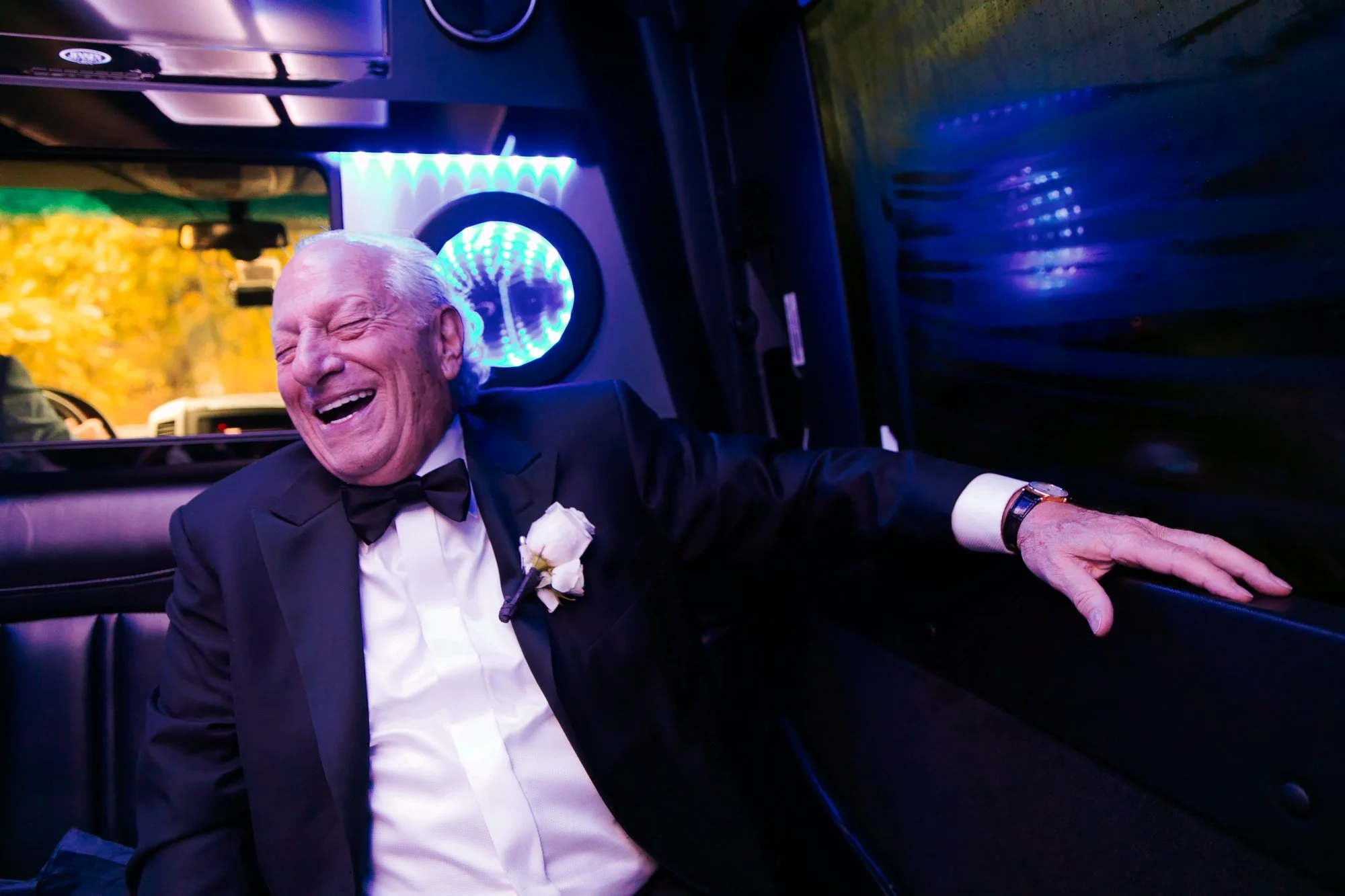 An elderly man in a tuxedo with a white dress shirt and a black bow tie, wearing a boutonniere, is sitting inside a limousine, laughing joyfully. The interior is dimly lit with blue and purple lights, and the window shows a blurred outdoor scene with