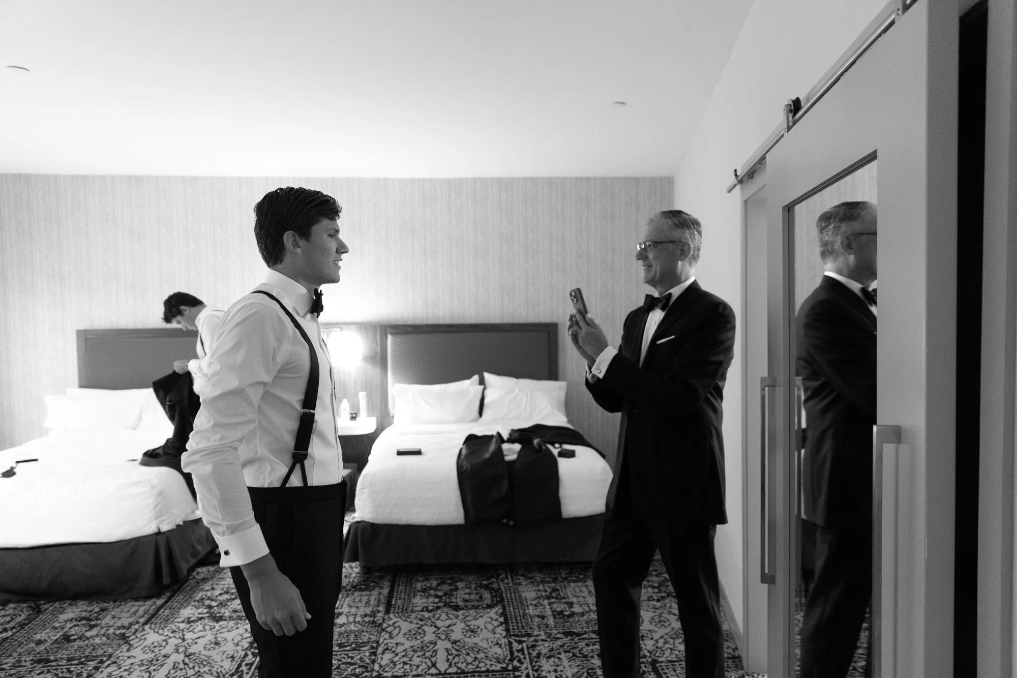 Three men dressed in tuxedos and formal attire inside a hotel room, with two beds and a mirror on the wall.