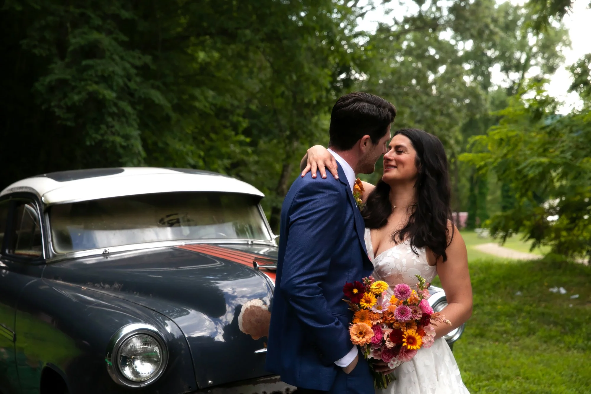 A newlywed couple embracing outside near a vintage car, surrounded by green trees, she holding a bouquet of colorful flowers.