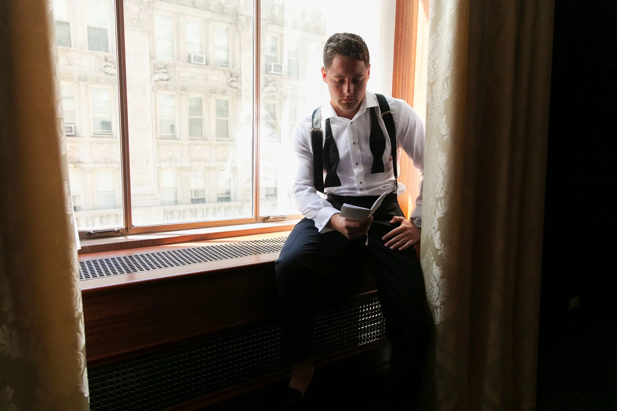 A young man in a tuxedo with a bow tie hanging around his neck is sitting on a window seat, looking at a small booklet, in a room with large window and curtains.