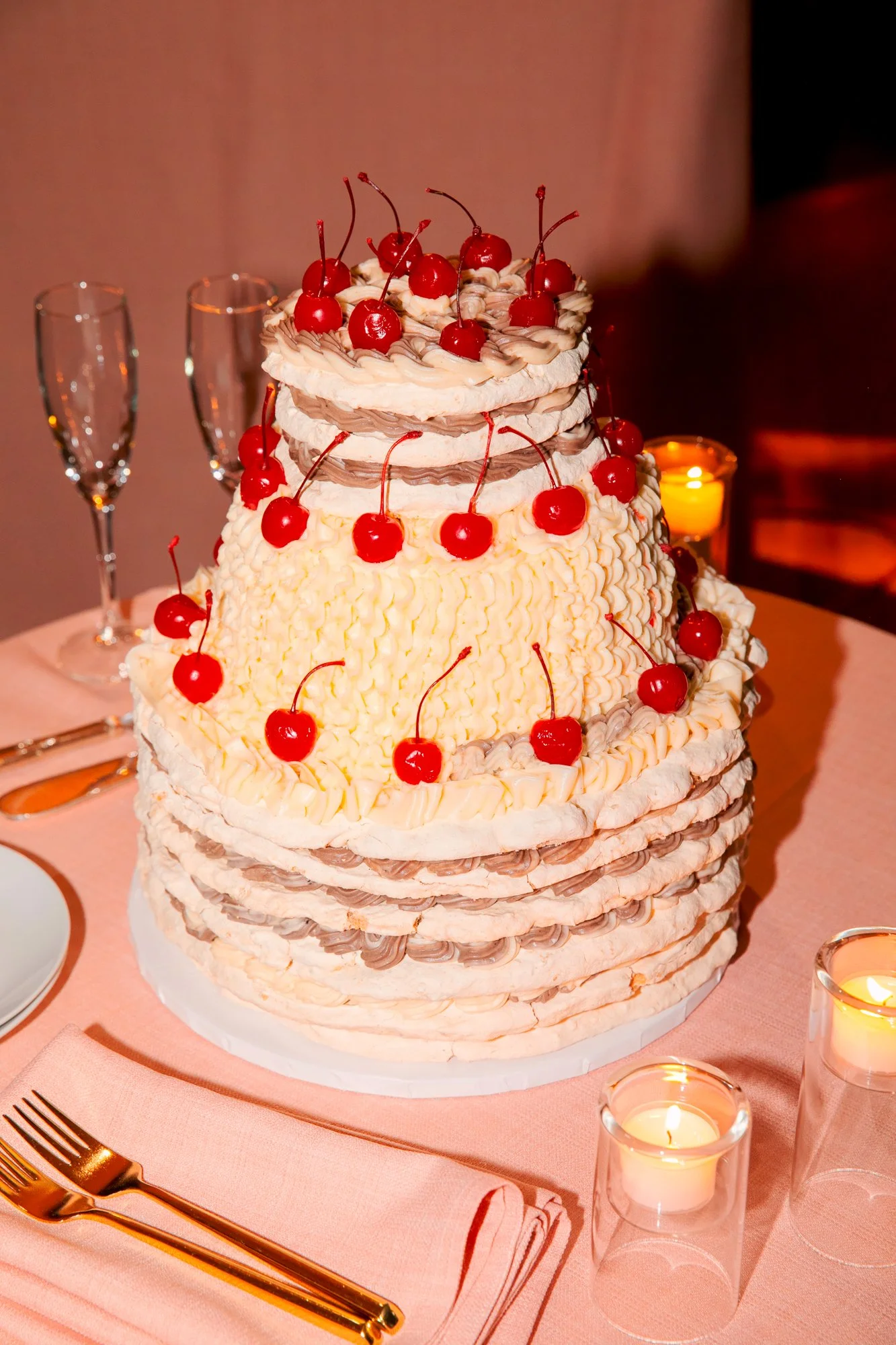 A tall multi-tiered cake decorated with cherries, surrounded by lit candles and set on a pink tablecloth with gold-colored cutlery.
