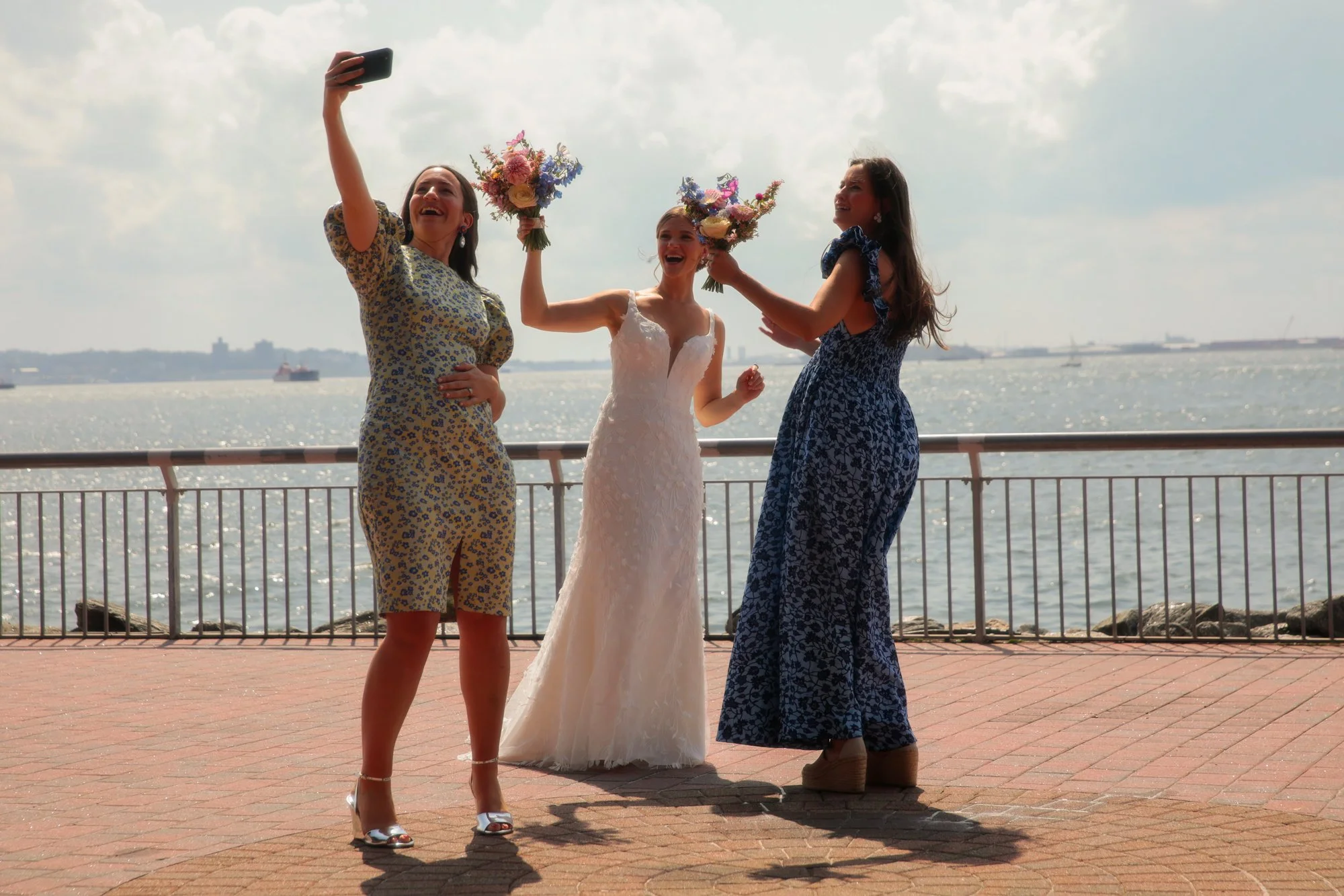 Three women standing by a waterfront, taking a selfie while the woman in the center, dressed in a wedding gown, holds a bouquet of flowers and the two women on either side also hold bouquets. The scene is sunny with ships in the distant water.