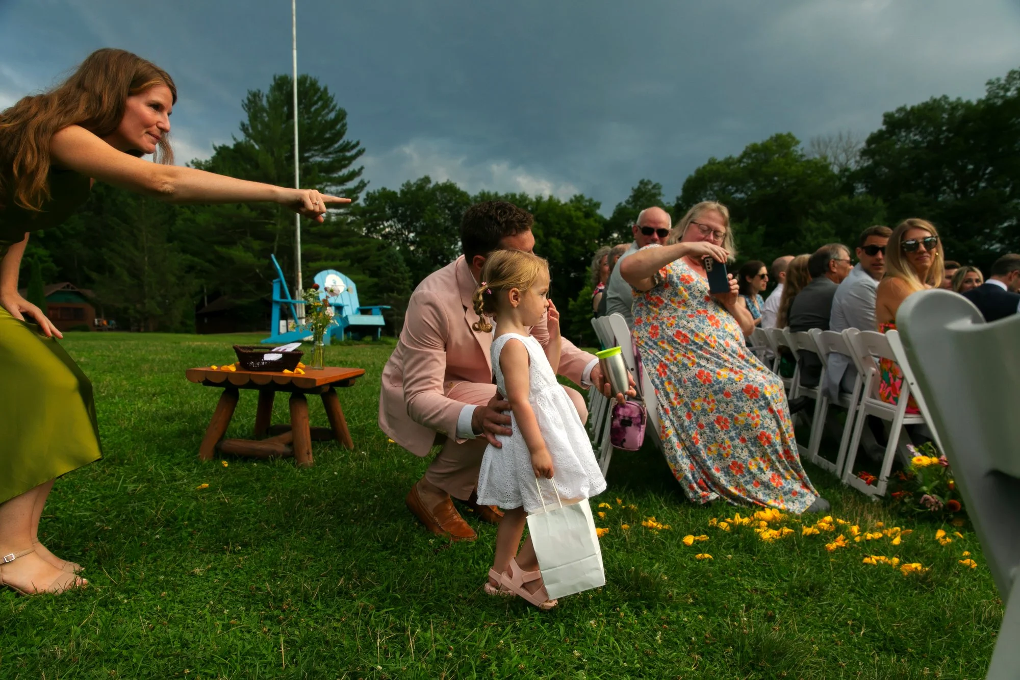 A woman is pointing towards a seated man and child at an outdoor event on a grassy area, with other seated guests and trees in the background.
