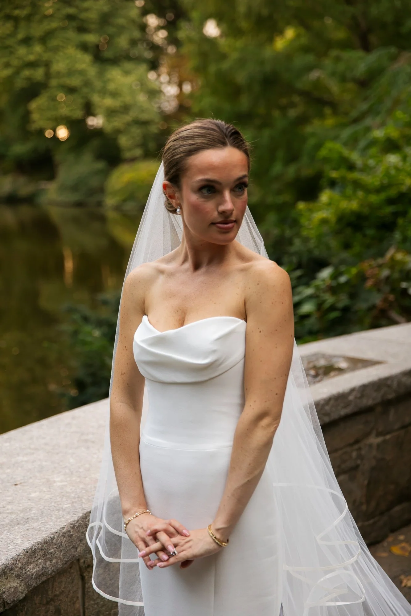 A bride standing outdoors in a white strapless wedding gown, wearing a veil and jewelry, with a natural, green background.