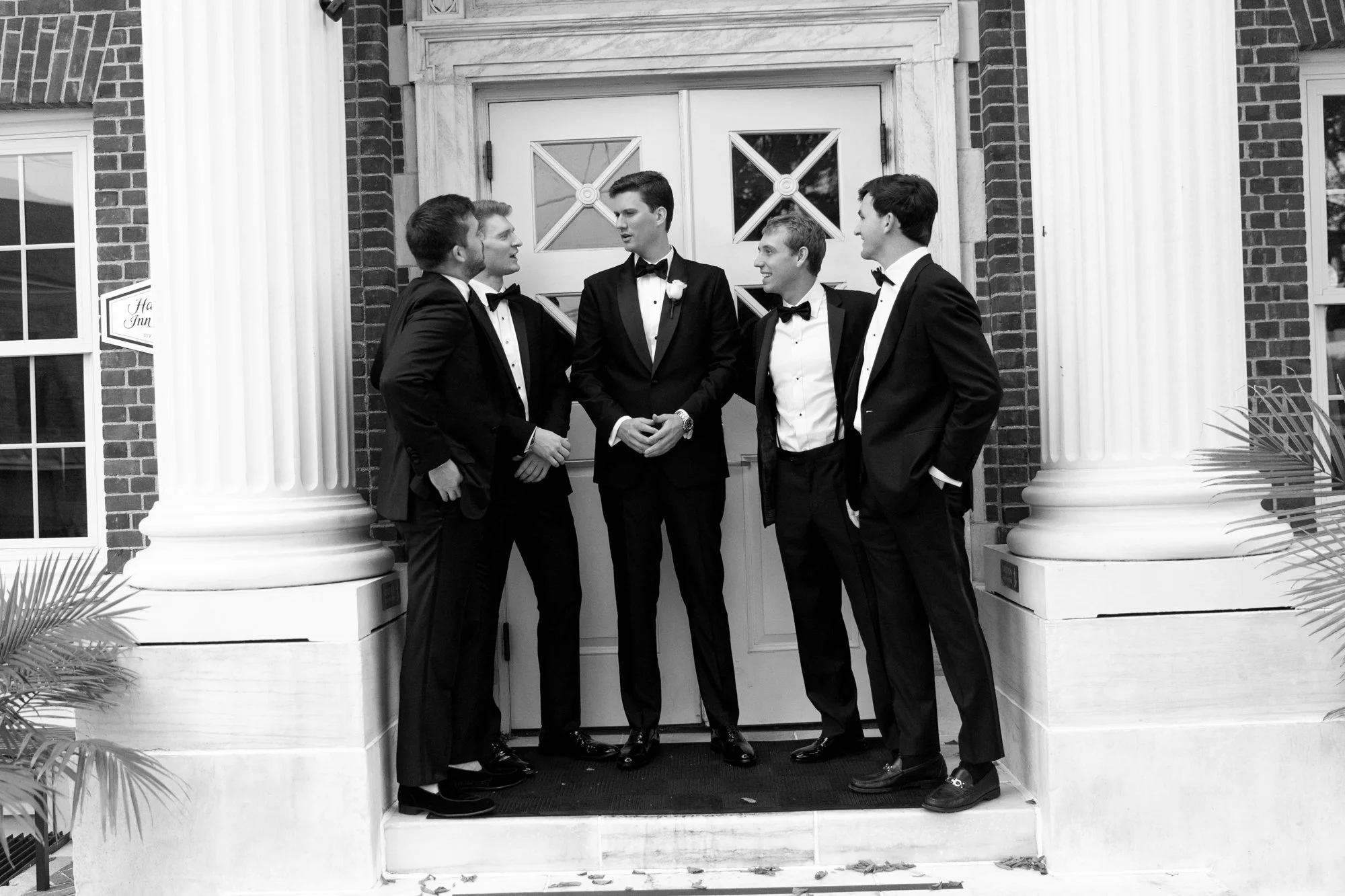 Five men dressed in black tuxedos and bow ties standing in front of a building with large white columns, engaged in conversation