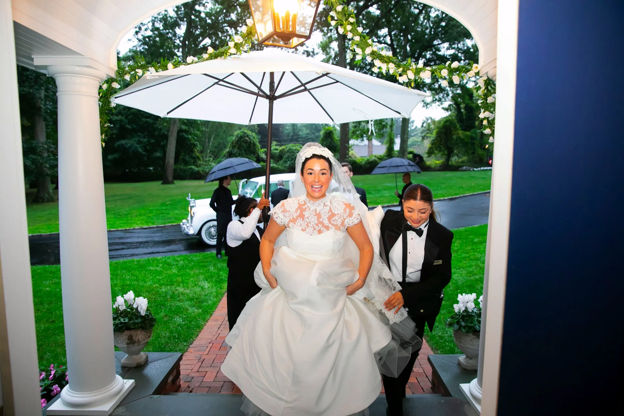 Bride in a wedding dress and veil stepping out of a house on a rainy day, smiling and holding her dress, with an umbrella held overhead. Two women in black tuxedos assist her. The background shows a green lawn, trees, and a white vintage car.