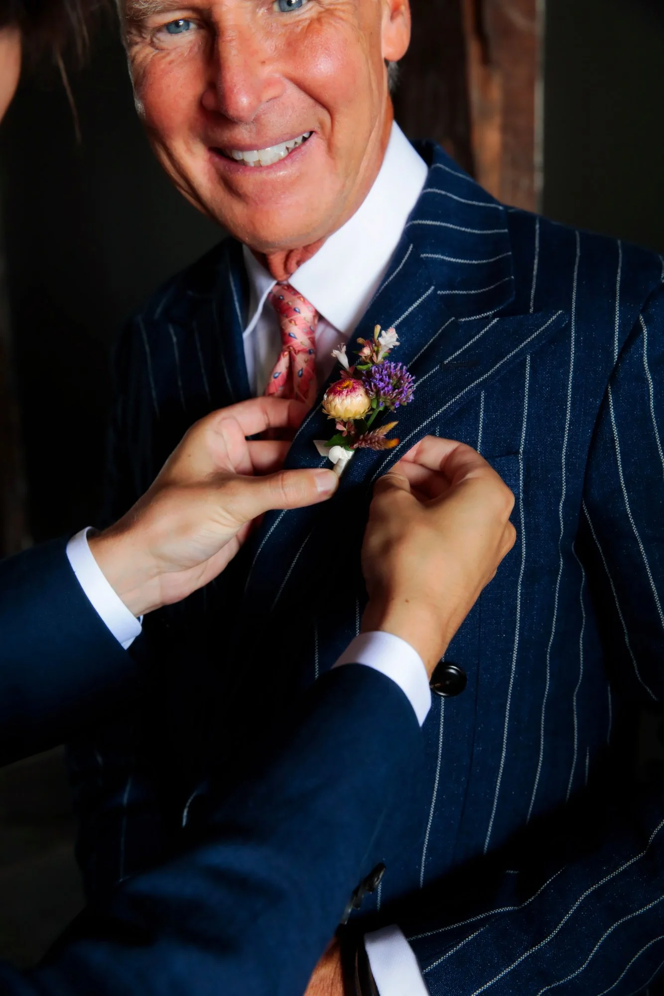 A man in a pinstripe suit is having a colorful flower boutonniere pinned to his lapel, smiling at the camera.