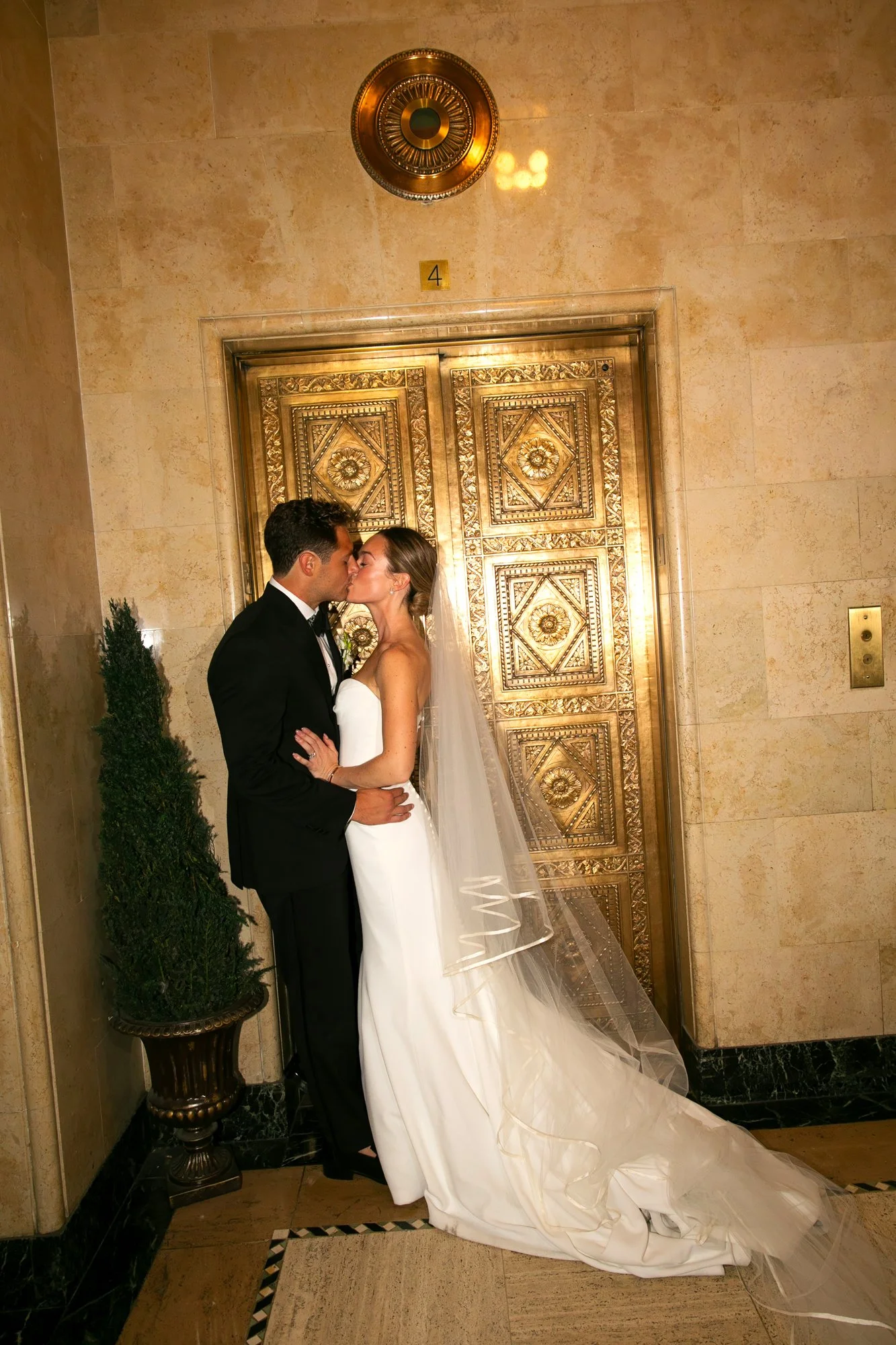 A bride and groom kissing in front of an ornate gold elevator door in an elegant setting.