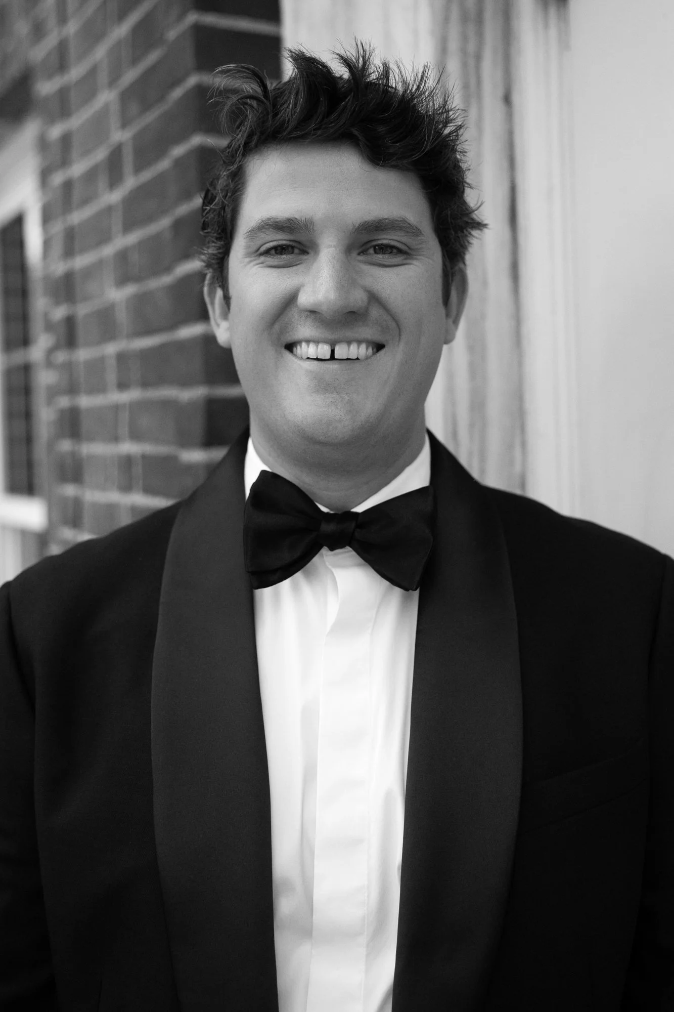 A smiling young man with dark messy hair wearing a tuxedo with a bow tie, standing outside near a brick wall.