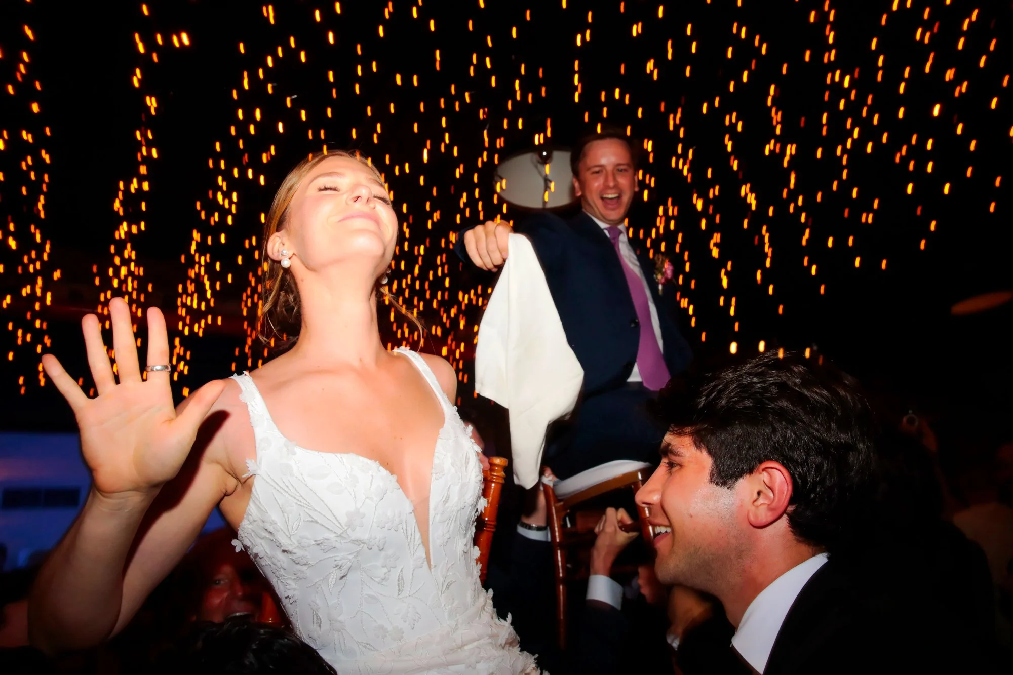Wedding reception with bride in a white dress with floral embroidery, raising her hand, and groom in a suit and tie, sitting on a chair with a joyful helper, under string lights in a dark setting.