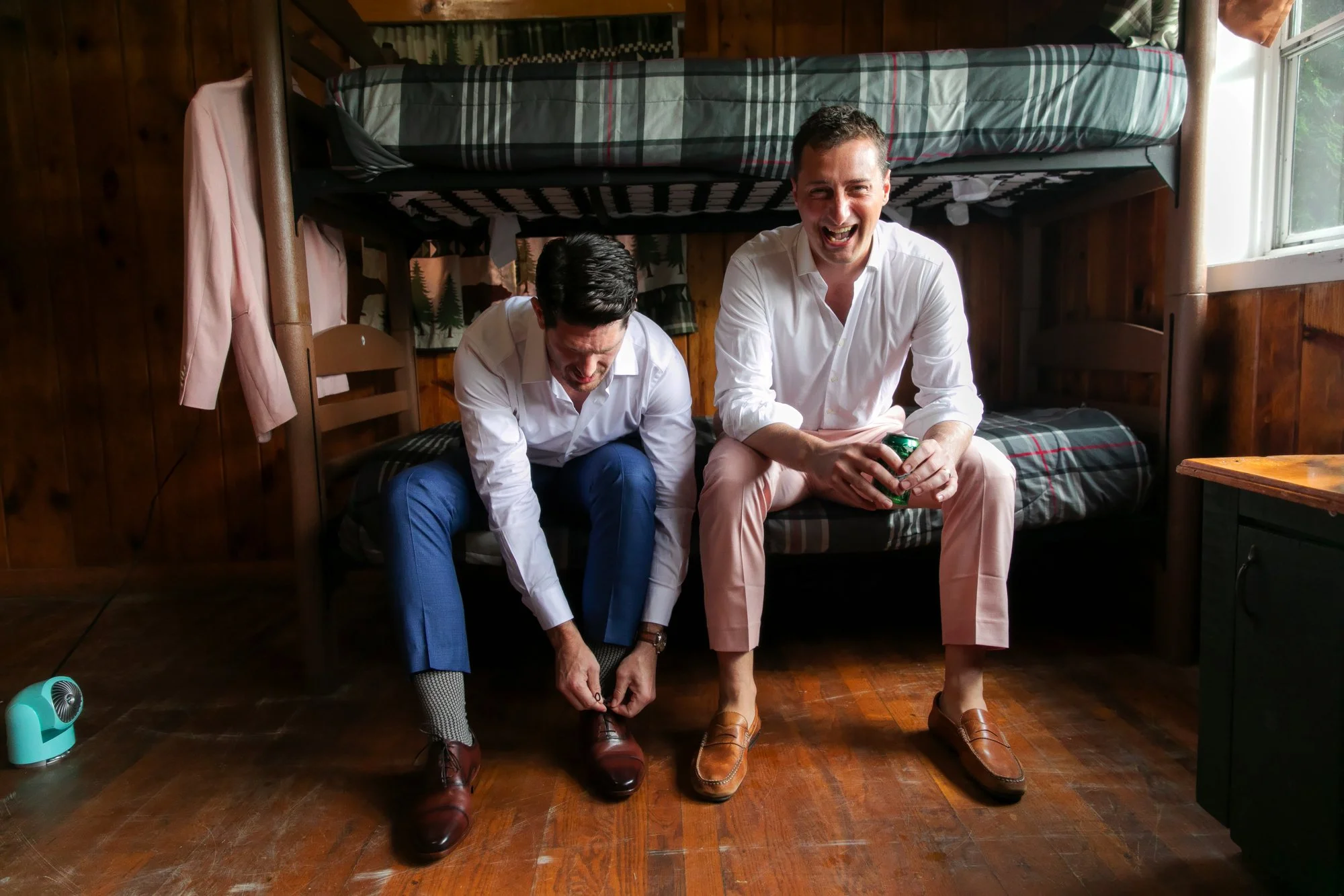 Two men in white shirts and dress shoes sitting in a cabin bedroom, one tying his shoe while the other is holding a soda can and laughing.