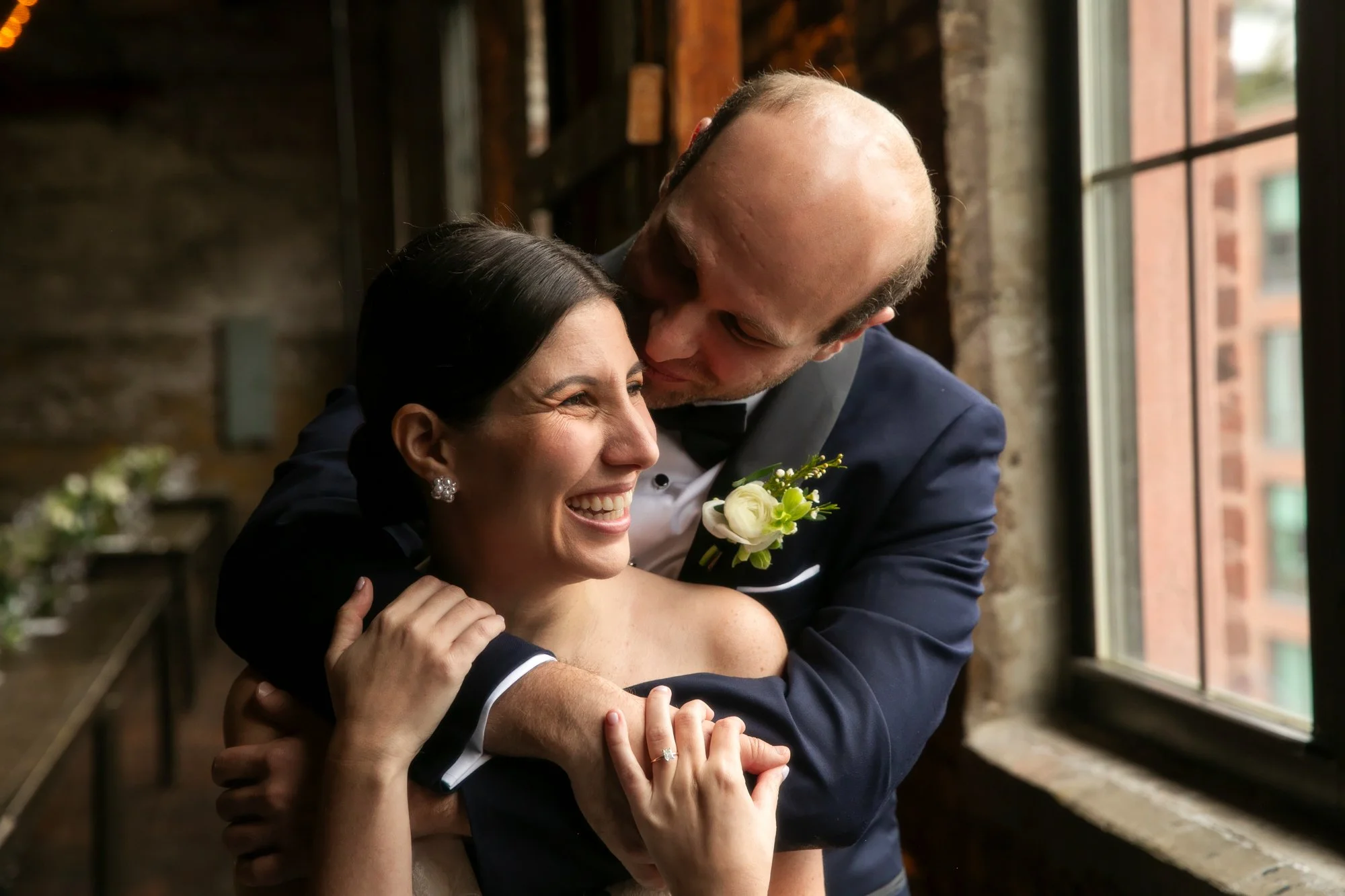 A couple on their wedding day, the groom with a tuxedo and boutonniere, embraces and kisses the bride, who is smiling and wearing earrings and a wedding ring, near a window with natural light.
