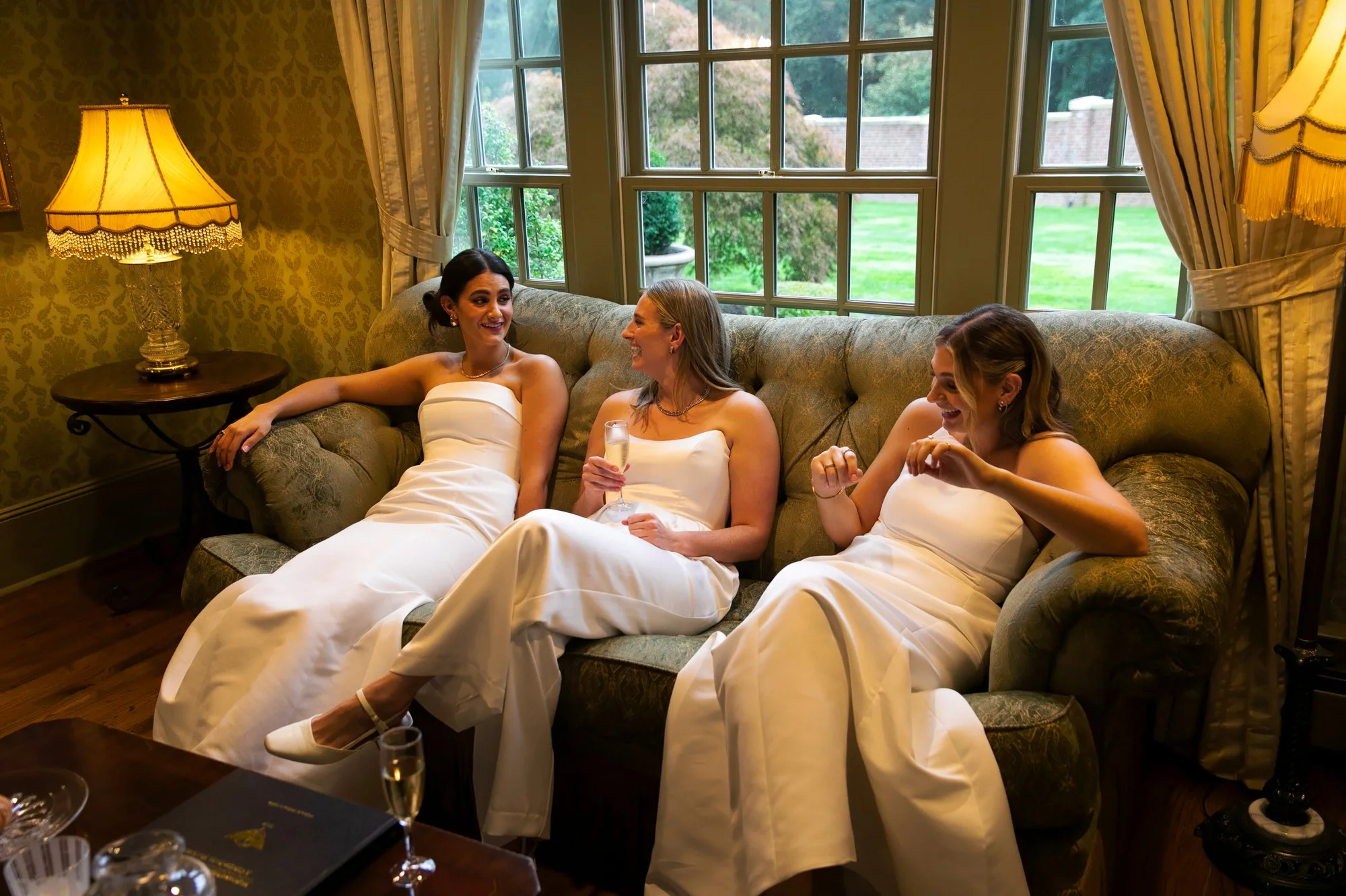 Three women in white bridesmaid dresses sitting on a vintage sofa, smiling and laughing indoors with a window showing a green lawn outside.