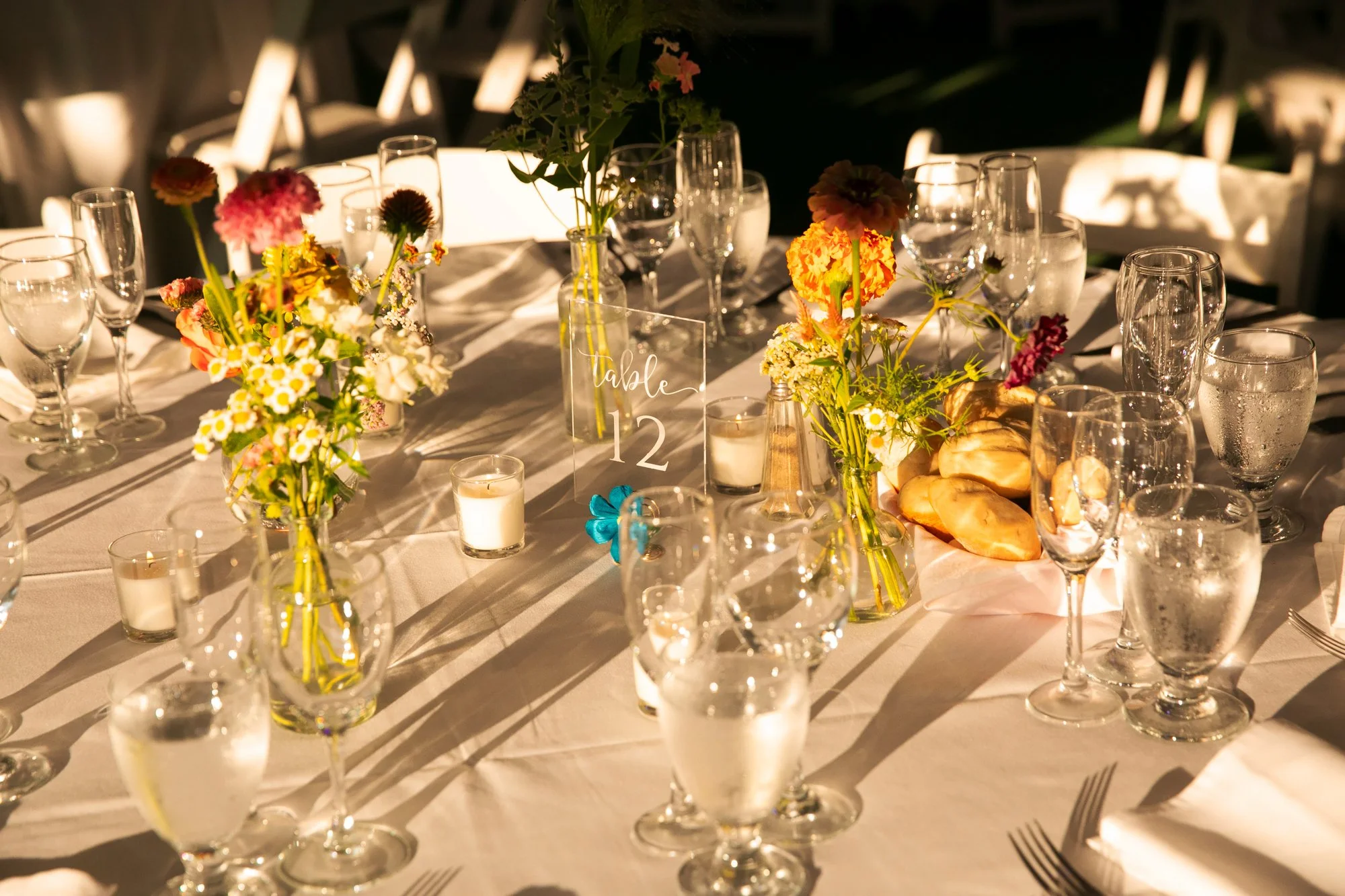 Elegant banquet table decorated with colorful flowers in glass vases, surrounded by glasses, candles, and tableware, with a table sign reading 'table 12'