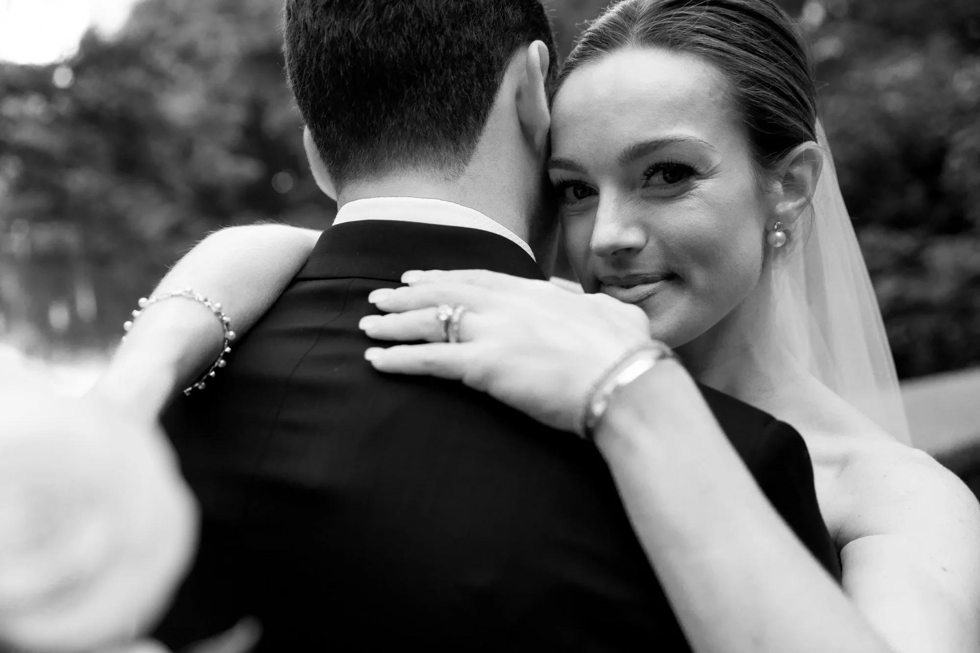 Black and white photo of a bride and groom hugging closely, with the bride smiling and looking at the camera. The bride's face is visible, with her hand resting on the groom's shoulder, showing her wedding ring and jewelry.