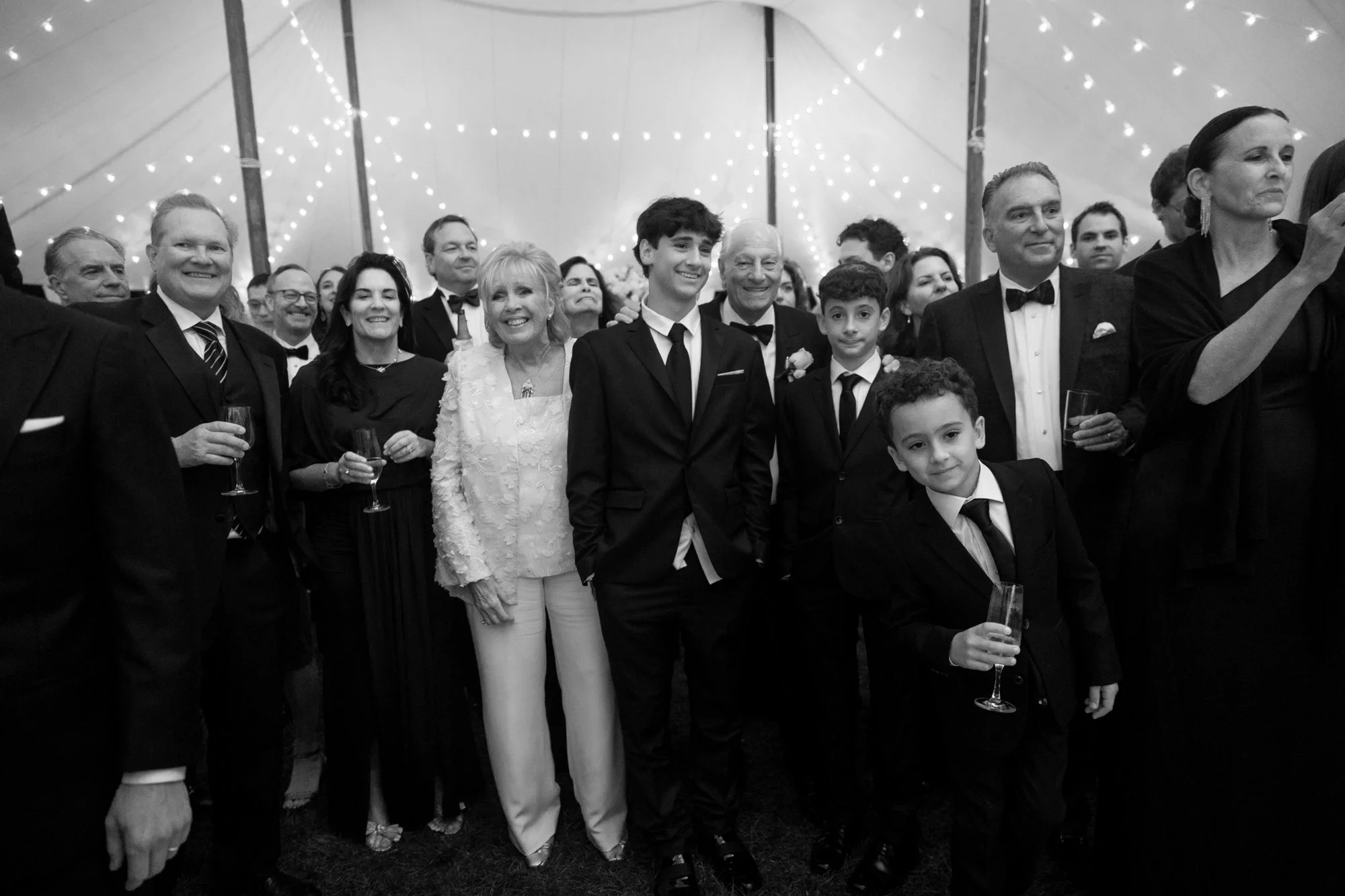 Group of people at a celebration event, dressed in formal attire, smiling and holding glasses, inside a decorated tent with string lights.